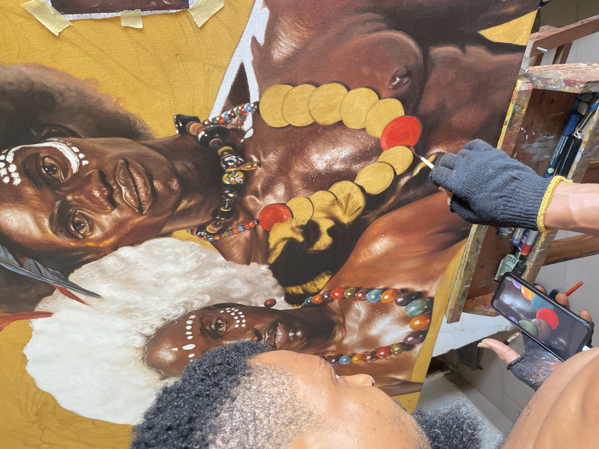 The image shows an artist working on a large-scale painting of two individuals adorned in what appears to be tribal or cultural ornaments. The artist is capturing elements on a phone and using it as reference. The setting appears to be inside an artist's studio, with visible art supplies and the unfinished painting. The individuals in the painting have dark skin and are wearing what appears to be face paint and beaded necklaces. The style of painting is realistic. The specific location within Ado, Nigeria, is indiscernible from the visual information available.