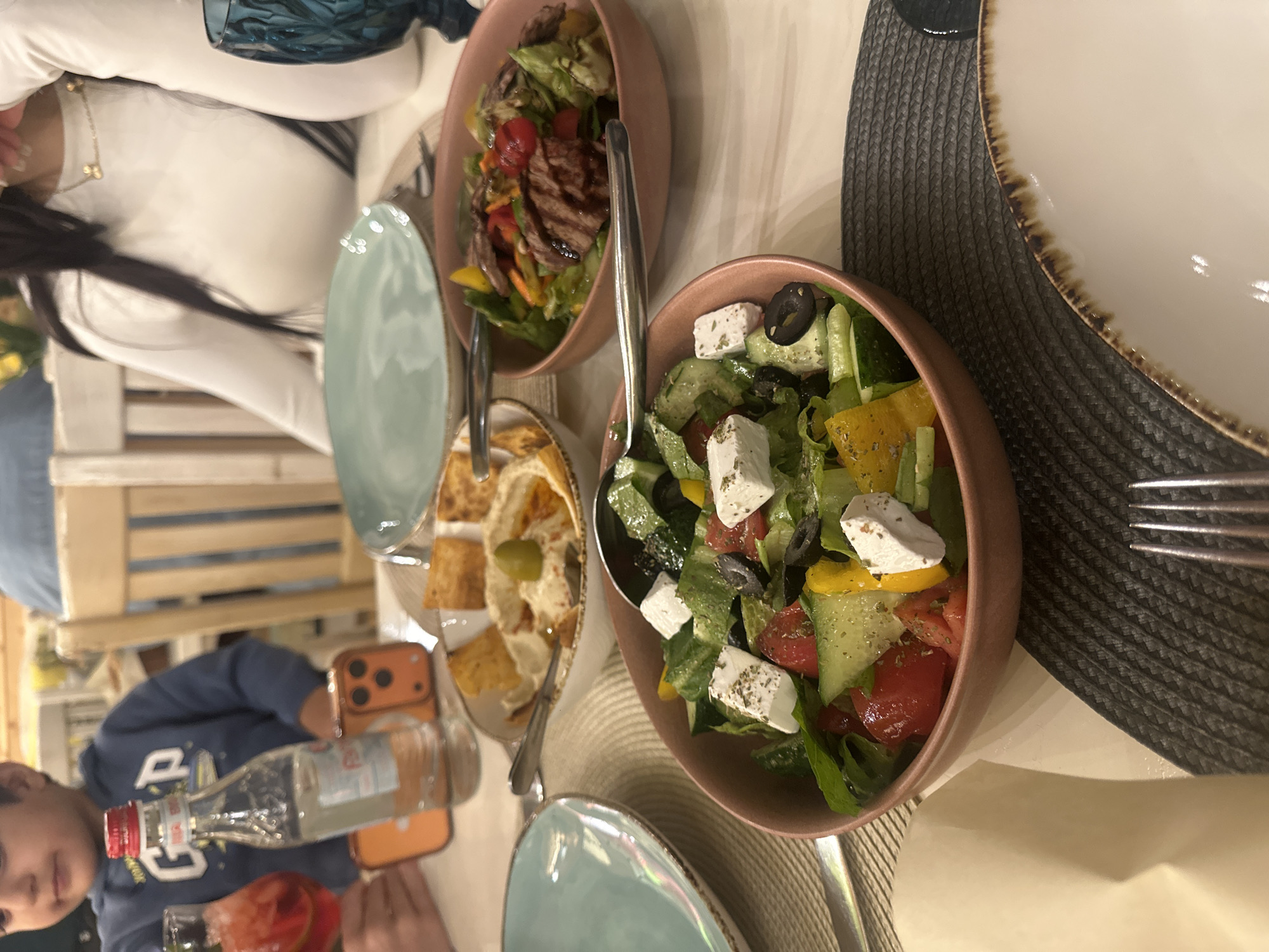 The image depicts a dining table laden with food. A bowl of salad with feta cheese, cucumbers, tomatoes, peppers, and black olives occupies the foreground. To its left is a hummus plate with pita bread. Another bowl contains a salad with grilled meat and vegetables. A plate, cutlery, a bottle of liquid, and an orange cell phone are also visible on the table. Two individuals are partially visible: one is wearing a white long-sleeved shirt and the other, a blue hoodie. The location is Tsakhkadzor, Armenia.