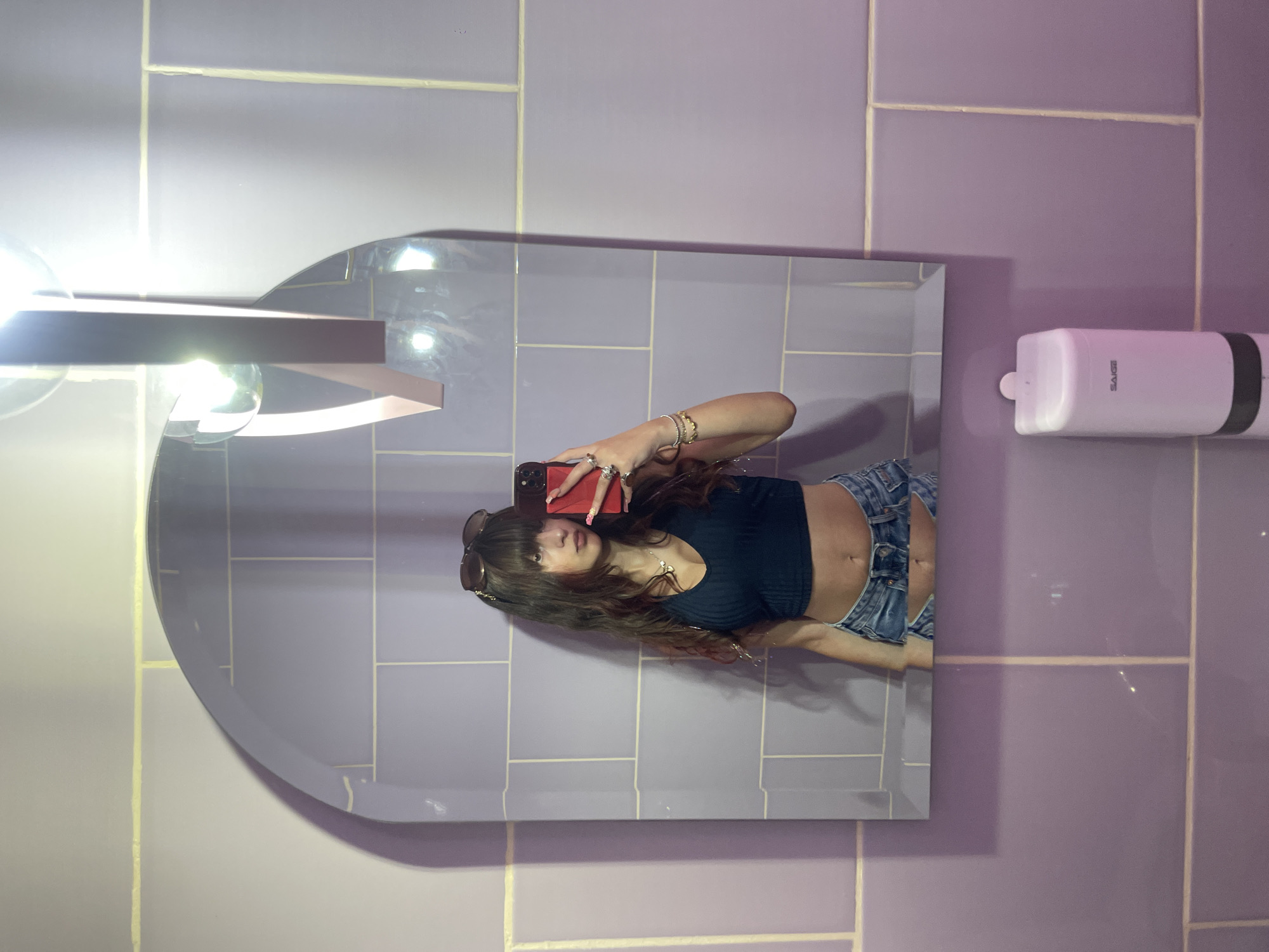 A young woman with long, dark hair and bangs is taking a selfie in a mirror. She is wearing a dark blue crop top and denim shorts. She has sunglasses pushed up on her head and is wearing rings and bracelets. The mirror is framed by light purple tiles. A soap dispenser is visible on the wall below the mirror. The scene appears to be in a public restroom.