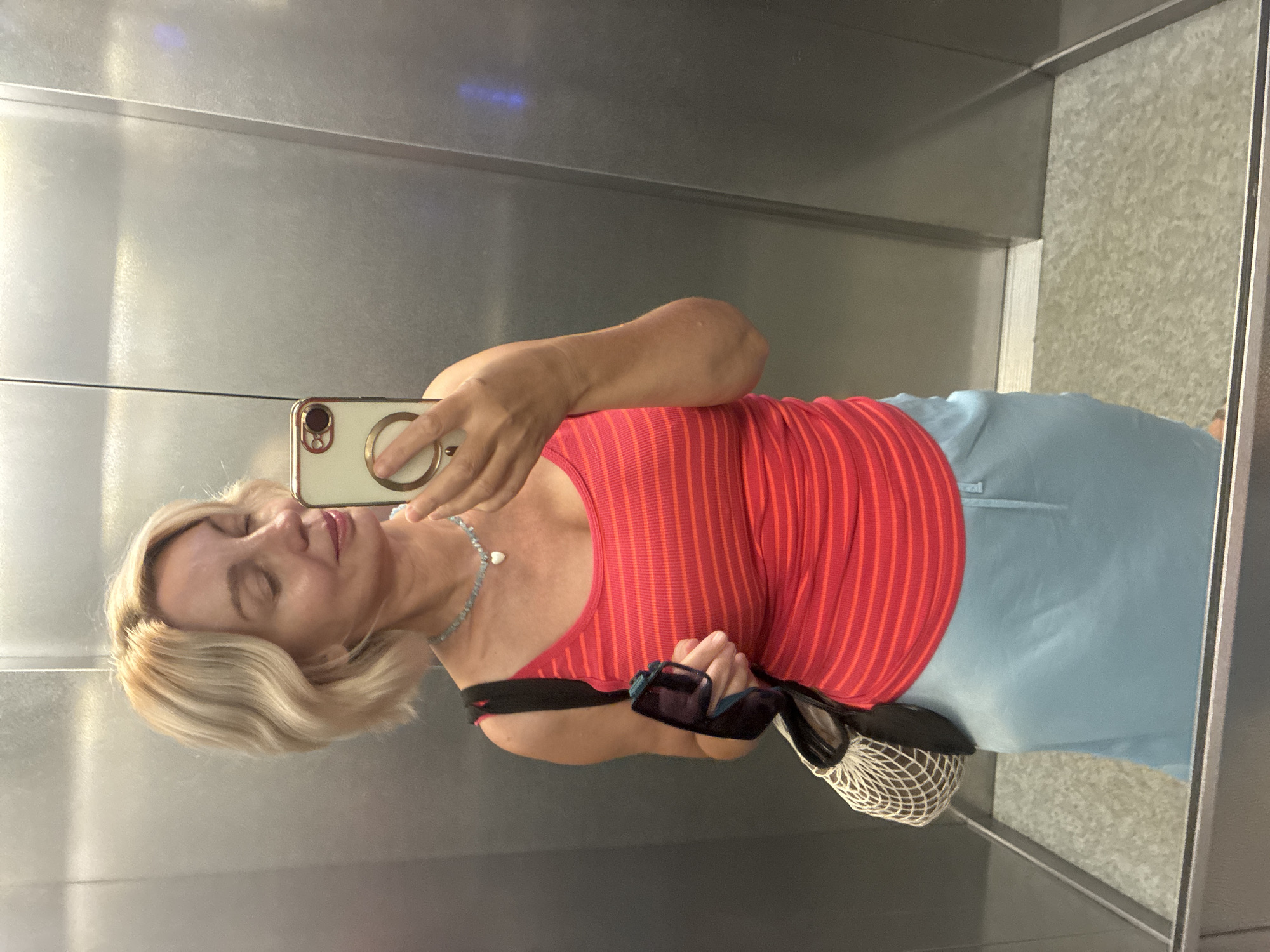 A woman with blonde hair is taking a selfie in an elevator. She is wearing a red ribbed tank top and light blue pants. She has on a necklace with a white heart-shaped pendant and is holding a pair of sunglasses. The elevator has brushed metal walls. The woman has her eyes closed and is smiling. She appears to be in good spirits. The lighting suggests it is daytime, and there are no visible clues about the weather. The setting is clearly an elevator, indicated by the reflective walls and the typical structure of an elevator interior. There is no visible text in the image.
