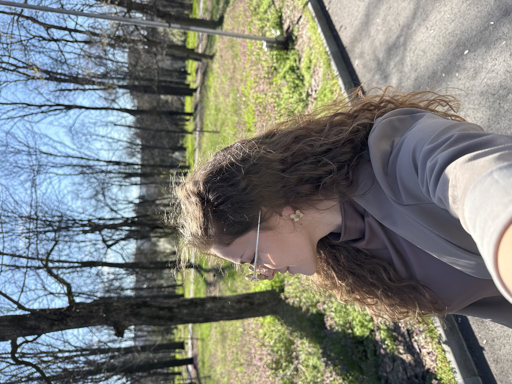 A medium shot captures a woman with curly brown hair, wearing a light gray blazer over a darker gray shirt, and eyeglasses with flower-shaped ornaments. She is positioned outdoors, facing slightly downward and to the left. In the background, a row of leafless trees lines a grassy area speckled with small purple flowers. A paved pathway is visible on the right edge of the frame. Based on prior knowledge, the location is Razgrad, Bulgaria, identifiable by its characteristic urban park features.