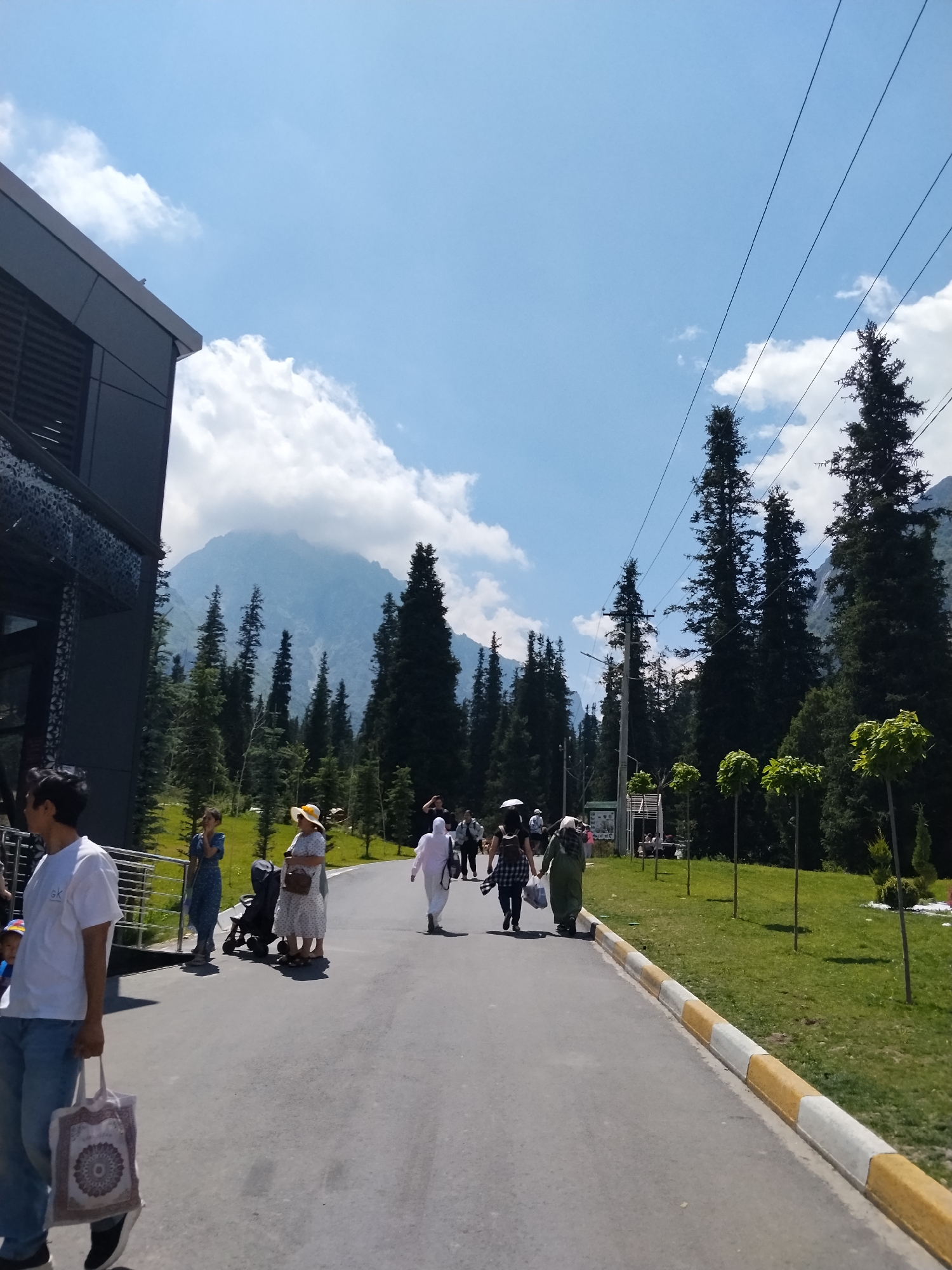 In Unknown, Kyrgyzstan, on a bright, sunny day, a group of people walk along a paved path lined with grass. A modern, dark-colored building with a patterned facade is on the left, and a mountain range, partially obscured by trees, rises in the background. Tall, dark green pine trees dominate the landscape, with manicured round trees planted along the right side of the path. Overhead, power lines stretch across the blue sky dotted with white clouds. Among the pedestrians, a man in a white t-shirt and jeans carries a tote bag. Further down the path, women in dresses and headscarves, some with a stroller, are walking. The scene depicts a leisurely outdoor activity in a mountainous, forested area.