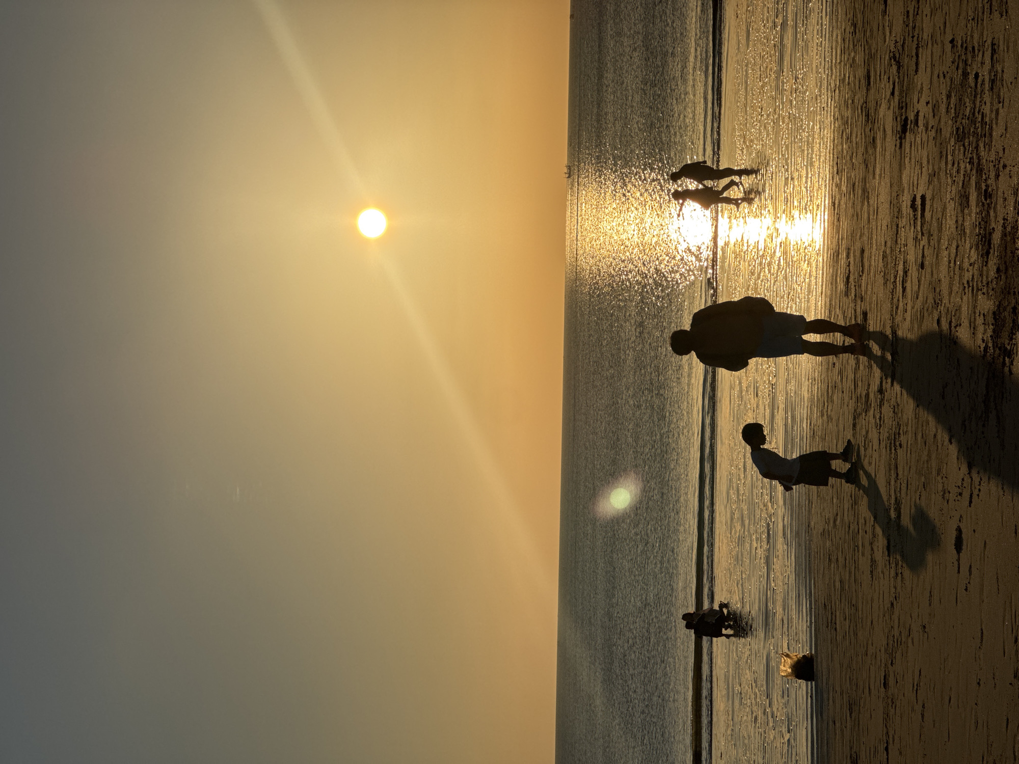The image captures a serene beach scene at sunset in Kuta Selatan, Indonesia. The sun, a bright orb, is low in the sky, casting a golden glow across the ocean and beach. The water is calm, with gentle waves lapping at the shore. Several people are visible on the beach and in the shallow water, silhouetted against the setting sun. In the foreground, a young boy is bent over, seemingly looking at something on the sand. Further back, a man stands with his back to the camera, casting a long shadow. Other figures are further out in the water, enjoying the evening light. The sky is a gradient of warm oranges and yellows, transitioning to a cooler blue at the top. The overall atmosphere is one of tranquility and natural beauty.