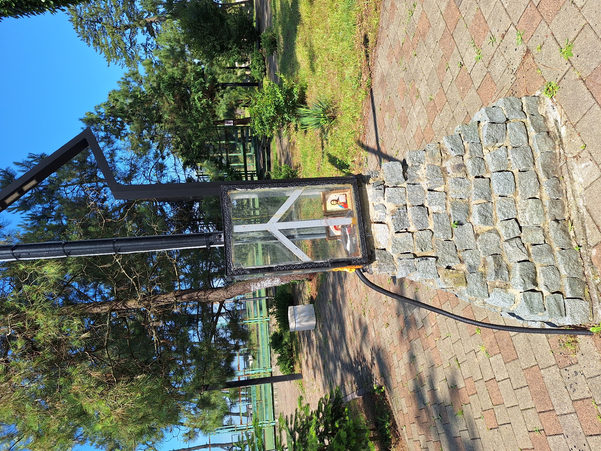 The image depicts an outdoor scene in Kobuleti, Georgia, featuring a stone pedestal topped with a black framed glass enclosure containing religious icons. A black corrugated hose runs from the enclosure down to the ground. The enclosure itself has a white peace symbol painted on its glass. Inside, two framed icons are visible: one of Saint George and the other of Jesus Christ. The pedestal is constructed from grey cobblestones. The surrounding environment includes a paved walkway made of interlocking bricks in shades of red and grey. Green grass and trees, including pine trees, are visible in the background. A light pole with integrated lighting fixtures is situated behind the pedestal. In the distance, a green fence, possibly bordering a sports field or park, can be seen. The sky is clear and blue.
