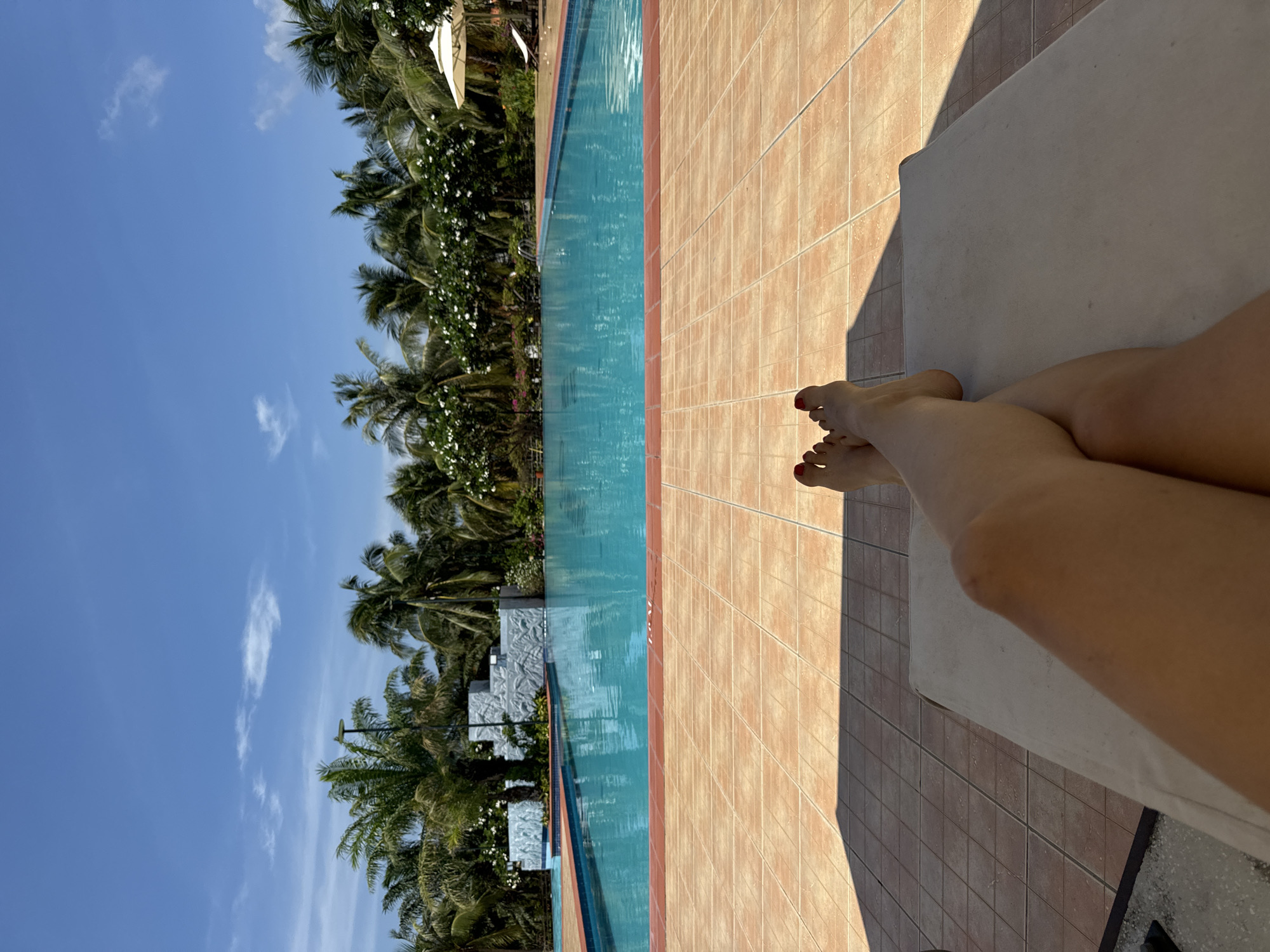 The image displays a first-person perspective of an individual's lower legs and feet, adorned with red toenail polish, resting crossed on a light-colored lounge chair positioned on a tiled surface. Strong shadows are cast onto the brown-orange tiled ground from the left, indicating direct sunlight. In the midground, a large, rectangular swimming pool with clear blue water and a red-brown coping is central. A white decorative structure with grey textured facades is situated within or adjacent to the pool on the left, surrounded by water. In the background, a dense line of tall green palm trees and white flowering foliage borders the pool area. A white sun umbrella and a white lounge chair are visible on the right side of the pool deck. The upper portion of the image reveals a clear blue sky with minimal scattered white clouds. A black light pole stands among the palm trees on the left. This scene is located in Phu Thuy Ward, Vietnam.