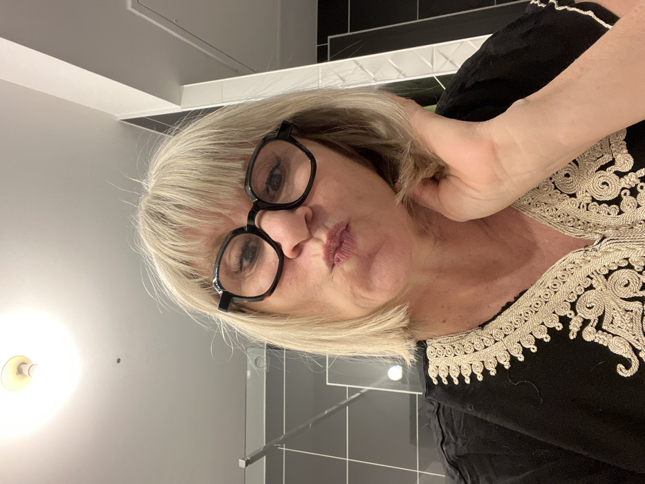 A selfie of a woman with blonde hair and black glasses pouting her lips. She's wearing a black top with intricate white embroidery along the neckline. Her hand is positioned near her neck. The background consists of a bathroom setting, including a light fixture, tiled walls, and a towel rack.