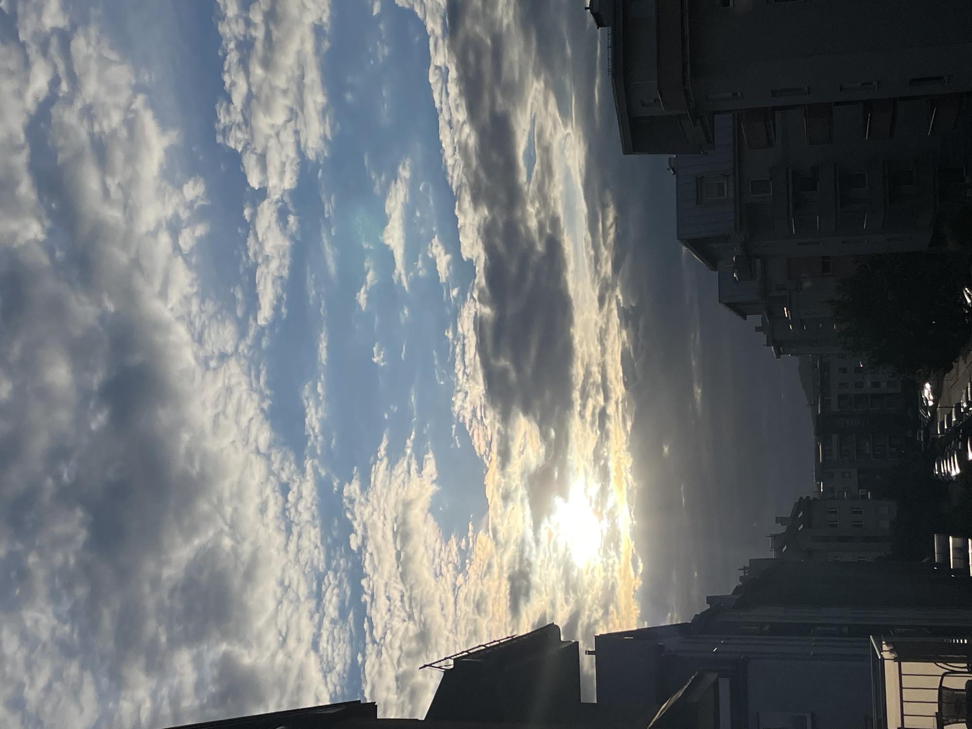 The image depicts a view of a city skyline with a dramatic sky filled with clouds. The sun is partially obscured by clouds, casting a bright glow and creating a luminous effect. The foreground features the side of a building with a balcony, and to the right, a row of apartment buildings is visible, with cars parked along the street below. The overall impression is a cityscape captured during either sunrise or sunset, with a focus on the interplay of light and shadow in the urban environment.