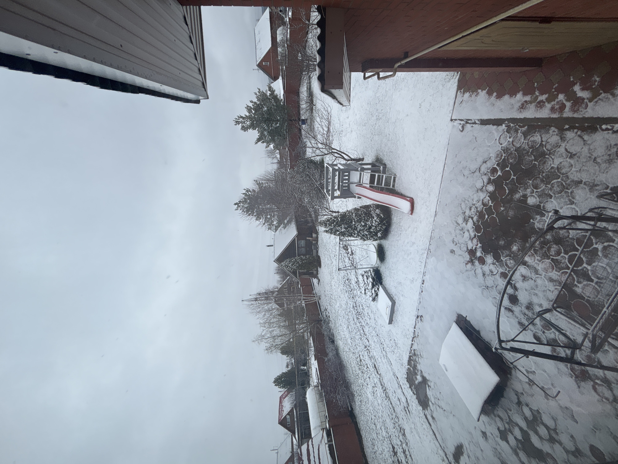The image shows a snow-covered backyard in Ферма, Russia. A wooden playset with a red slide sits in the middle of the yard. The ground is covered in a thin layer of snow, with visible footprints and patterns suggesting textured paving beneath. A metal chair sits to the right, partially covered in snow. Houses with snow-dusted roofs are visible in the background, and bare trees line the perimeter. The sky is overcast. The image is taken from an elevated position, possibly inside a house, looking out through a window. A portion of a building’s roof or overhang is visible in the upper left corner.