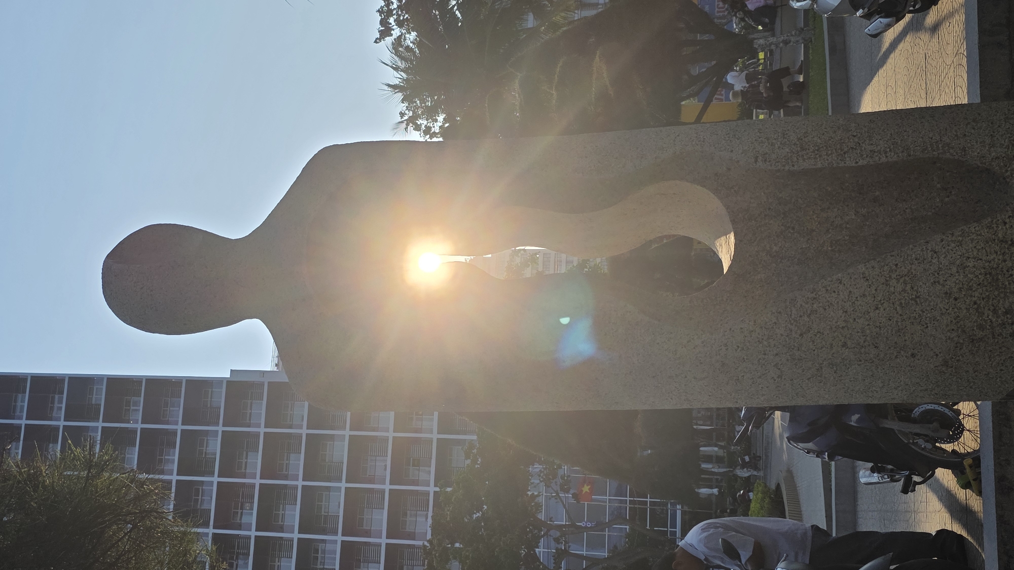 The image captures a bright sunny day in Nha Trang, Vietnam. The sun is directly behind a stone sculpture in the shape of a person with a hollow center. The sunlight is flaring around the edge of the statue. The buildings, trees, and people are visible through the statue, but the sun causes the image to be overexposed. The statue has a head and torso with a hole in the center. The architecture is more modern and boxy. There is a line of people on the sidewalk, and motorcycles and trees can be seen on the side of the image.