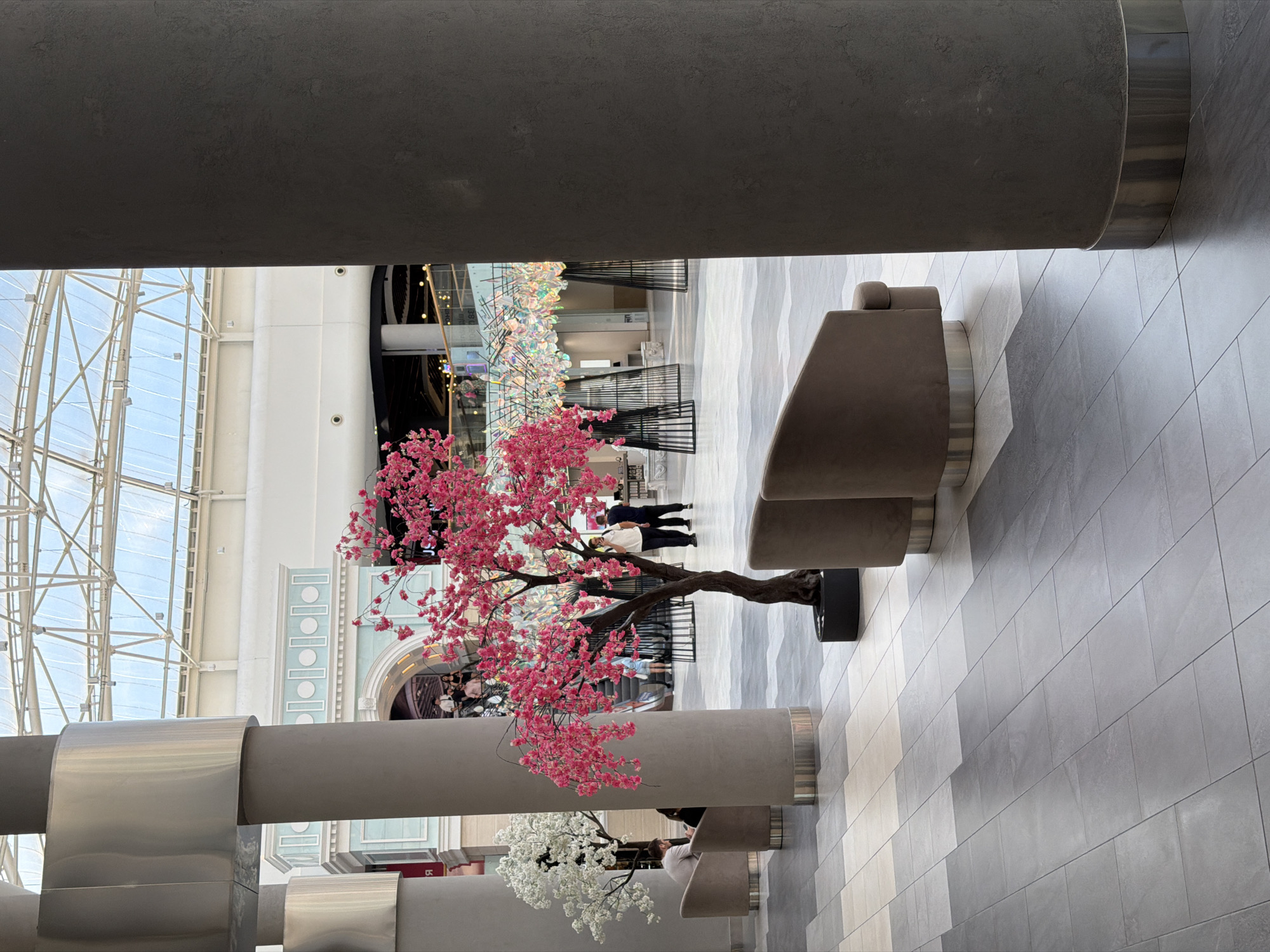 In an indoor shopping mall or public atrium, a decorative artificial cherry blossom tree with vibrant pink flowers is a central subject. Next to it, a similar tree with white blossoms is visible. The scene is characterized by large, gray cylindrical pillars and a polished gray tiled floor arranged in a geometric pattern. A modern, beige upholstered bench or seating area is also present.
In the background, a glass ceiling structure allows natural light to illuminate the space. There are glimpses of other shoppers and architectural elements, including an arched entrance and decorative vertical panels with circles. The overall atmosphere appears calm and modern. The time of day is indeterminate due to the artificial lighting and glass ceiling, and the weather is irrelevant as the setting is indoors. No visible text is discernable. The location context is Kyiv, Ukraine.