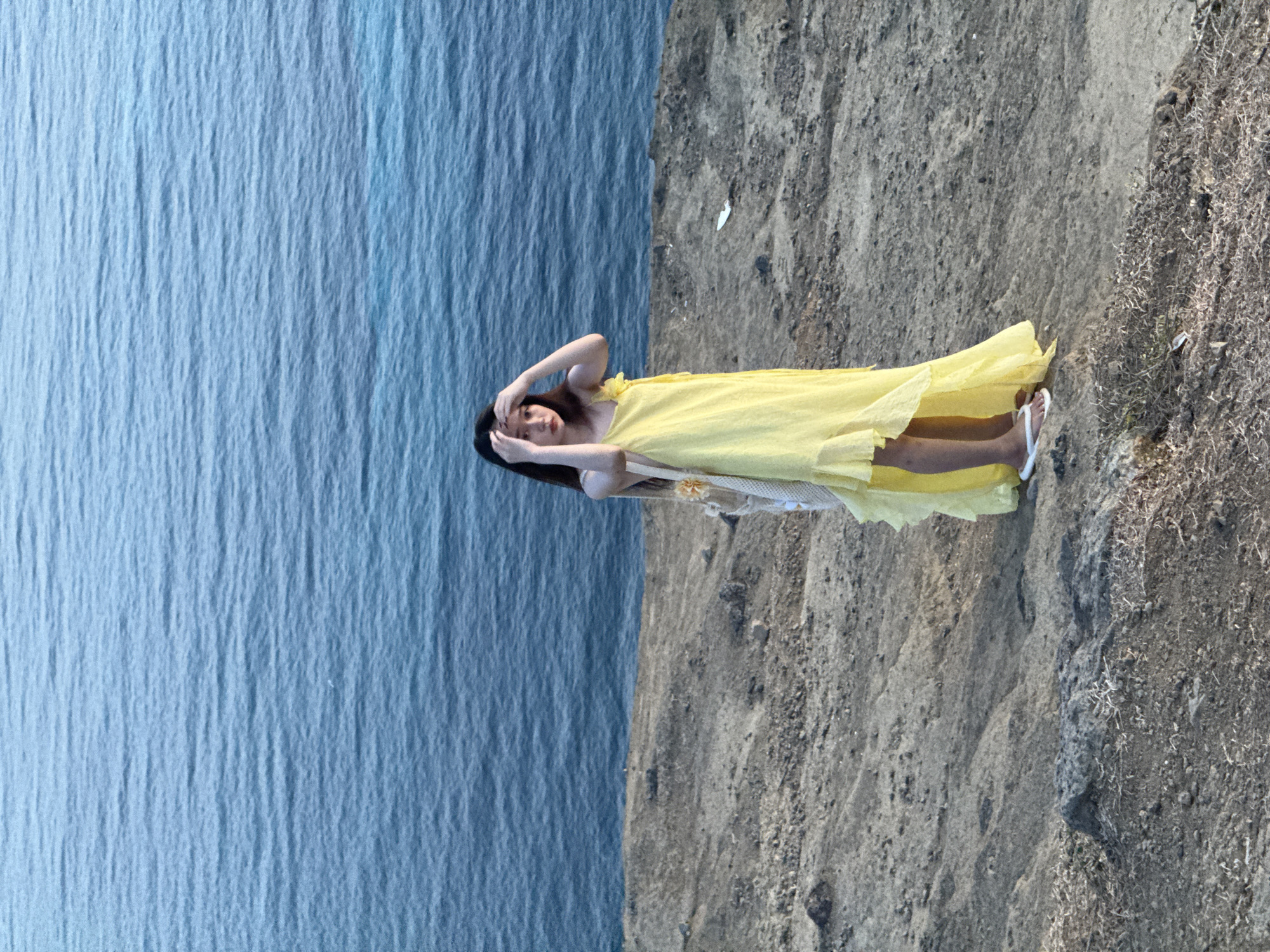 A young woman stands on a rocky, arid hillside overlooking a vast expanse of blue ocean. She is wearing a flowing, bright yellow maxi dress with thin straps and a ruffled hem. Her long, dark hair is styled down, and she is touching her temples with both hands. A woven tote bag with a floral embellishment hangs from her left shoulder. She is wearing white flip-flops. The background is dominated by the textured, choppy surface of the ocean, which occupies the upper half of the image. The ground beneath her is a mix of dry earth, small stones, and sparse, dry vegetation. The scene appears to be set in a coastal, natural environment, likely on Lý Sơn Island, which is part of the Lý Sơn Special Administrative Region in Vietnam.