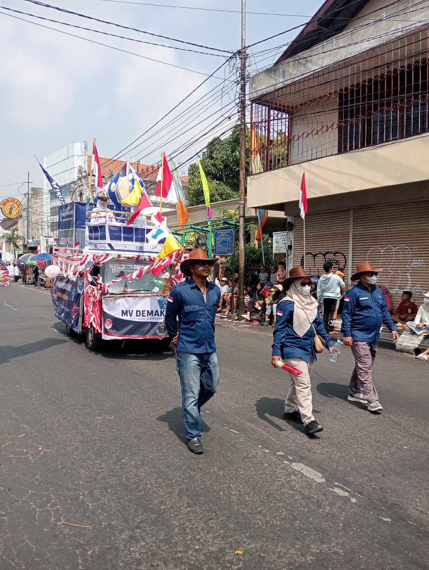 This image depicts a street parade in Bintoro, Indonesia. A large, decorated float resembling a ship, labeled "MV DEMAK," is prominently featured in the center of the road. The float is adorned with numerous flags, banners, and red and white streamers. Several people are visible on and around the float, some appearing to be participants or organizers.
In the foreground, three individuals are walking abreast on the right side of the road. The person on the left is a man wearing a blue long-sleeved shirt, jeans, and a brown cowboy hat. He has white markings on his face. The middle person is a woman wearing a blue long-sleeved shirt, tan pants, a cream-colored hijab, and a mask. She is holding a red baton in her right hand and a water bottle in her left. The person on the right is a man in a blue long-sleeved shirt, tan pants, and a brown cowboy hat, wearing a mask.
The street is lined with spectators sitting on the curb and standing along the sidewalk, observing the parade. Buildings with barred windows and awnings are visible along the right side of the street. Overhead wires are strung across the scene, indicating an urban environment. The sky is bright and appears to be daytime. The overall atmosphere suggests a festive community event.