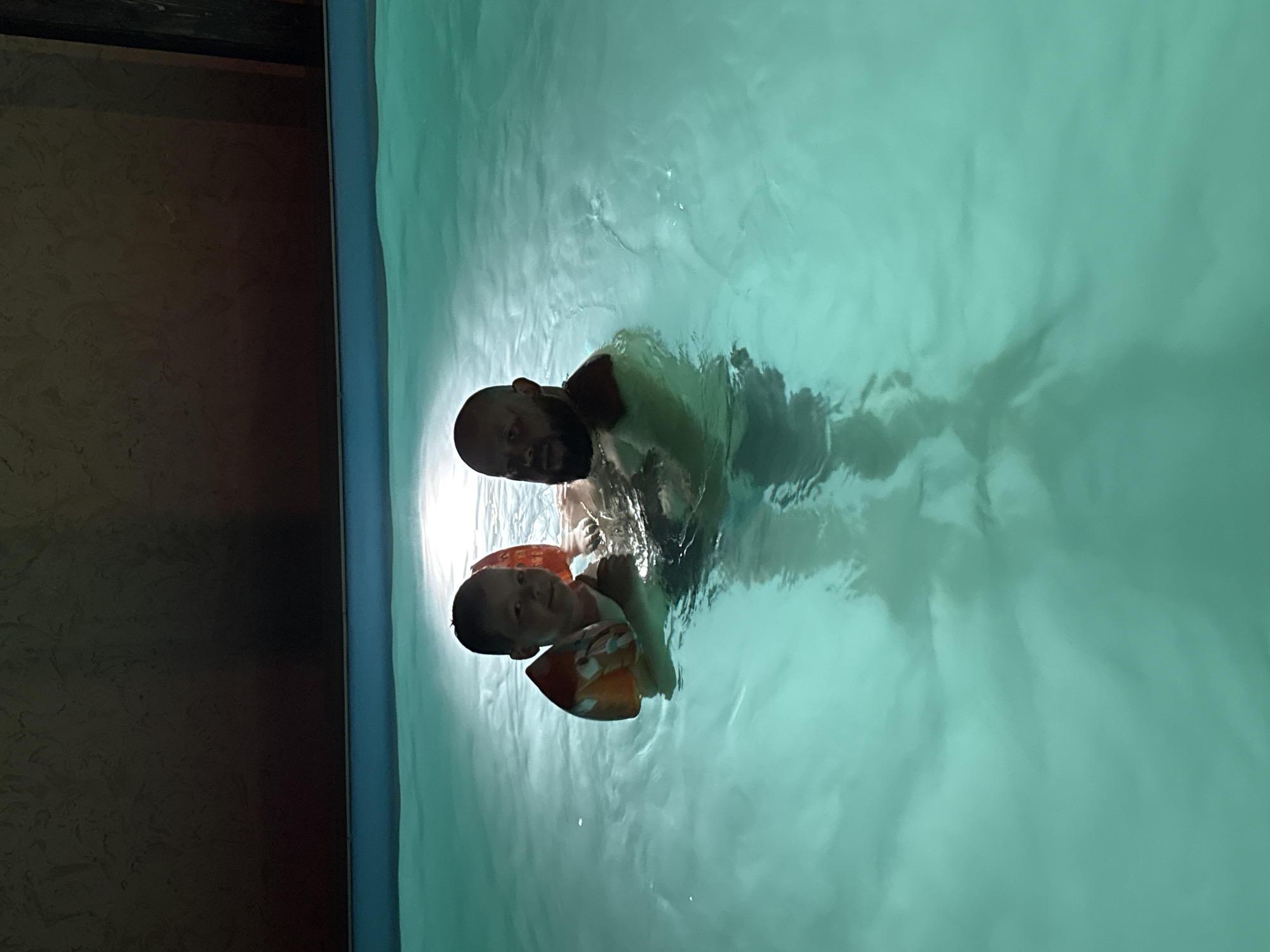 A man and child are in a body of water, possibly a pool. The child is wearing orange arm floaties. The man is holding the child. The water is a pale blue/green color. The location is unspecified, not identifiable as Krasnaya Polyana, Russia, from visual evidence in the image.