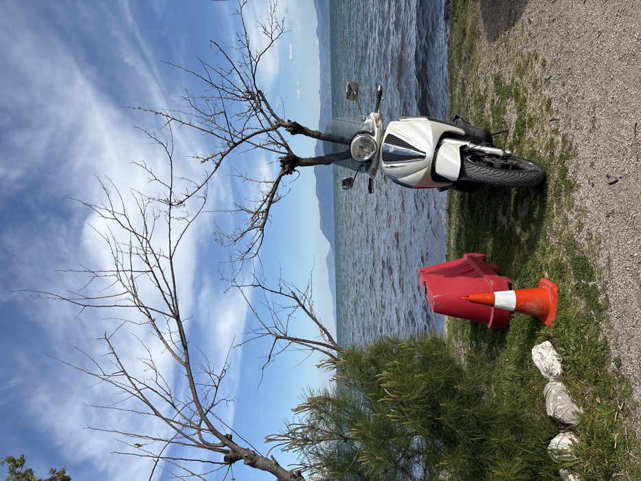 The image shows a white scooter parked on the shore of a body of water with a blue sky and white clouds above. The scooter is parked in the middle of the image facing the water. To the left of the scooter is a red and white traffic cone, and to the right of the scooter is a red plastic barricade. There are trees in the background and the water is slightly choppy. The image was taken in Vasiliki, Greece, at latitude 38.905 and longitude 22.599. It is a clear, sunny day and the water is calm. The environment is a relaxed, outdoor setting with a focus on the scooter and its surroundings.