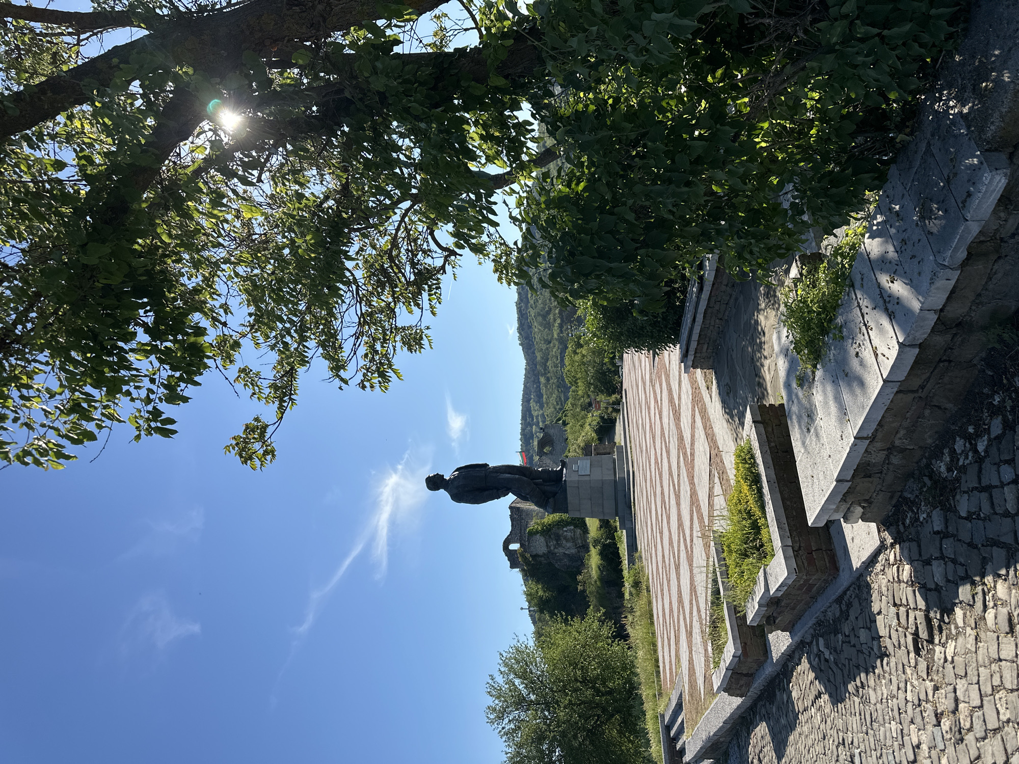 The image shows a bronze statue of a man in Lovech, Bulgaria. The statue stands on a tiled platform with a stone wall bordering a cobblestone street. A tree with green leaves overhangs the scene from the top left. In the background, there is a landscape with green vegetation and a partly ruined stone structure. The sky is blue with some wispy clouds. The sun is visible through the leaves of the tree, creating a lens flare effect.