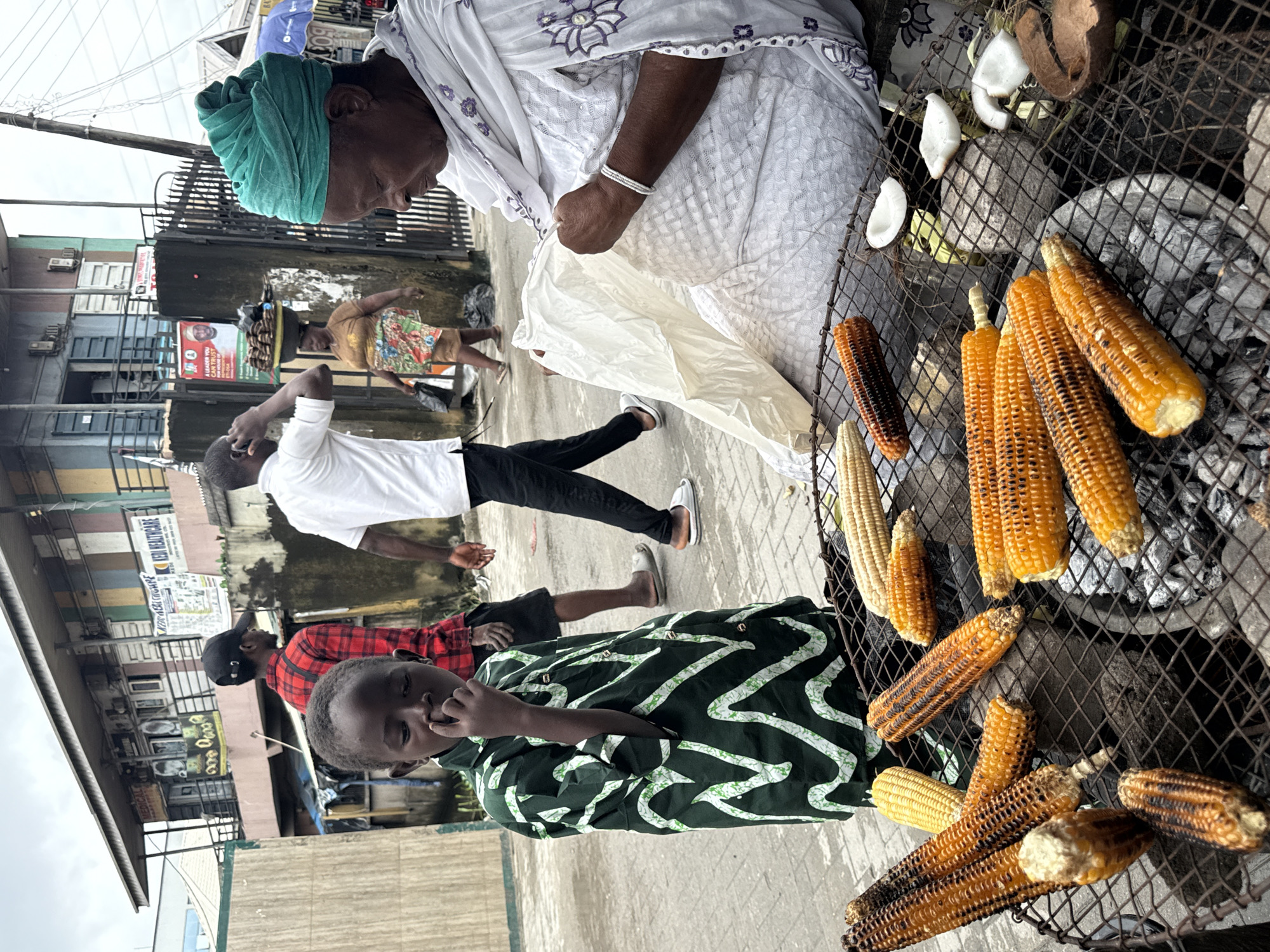 In Ado, Nigeria, an outdoor scene unfolds with a focus on a charcoal grill laden with corn. Several ears of corn, some visibly charred from roasting, are arranged on a metal grate. Alongside the corn, pieces of cracked coconut and what appears to be coconut shell are scattered.
In the foreground, a young boy in a green and white patterned shirt stands with his hand to his nose. To the right, an older woman, draped in a white garment and a vibrant green headwrap, sits near the grill, holding a white plastic bag. Her arm rests on the grill, and she appears to be engaged with the bag's contents.
In the background, people are walking past. One man in a red and black checkered shirt walks away from the viewer. Another man in a white t-shirt and black pants is also visible. Behind them, a building with a covered walkway and various signs is present, suggesting a commercial area. The overall setting indicates a street-side vending operation.
