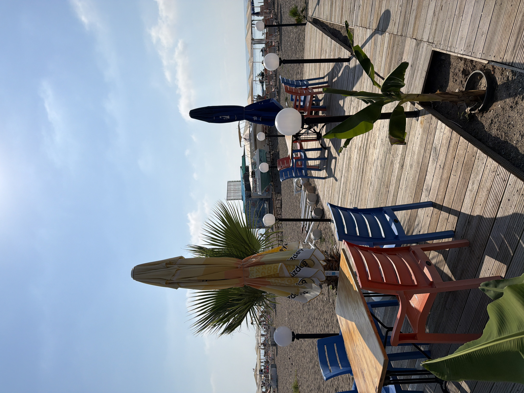 This image captures an outdoor seating area at a beachside establishment in Kemer, Turkey. The foreground features a wooden deck with a table and chairs, including blue and orange plastic chairs and a wooden table. Two large, closed umbrellas stand nearby, one yellow and white patterned and the other dark blue. Palm fronds are visible in the lower left corner, and a small banana tree is planted in a rectangular cutout in the deck to the right. Beyond the deck, a sandy beach is dotted with numerous beach chairs and umbrellas, with people lounging and enjoying the seaside. Several boats are docked in the shallow water, and the turquoise Mediterranean Sea stretches to the horizon under a bright blue sky with scattered white clouds. The scene conveys a relaxed atmosphere, typical of a coastal resort town.