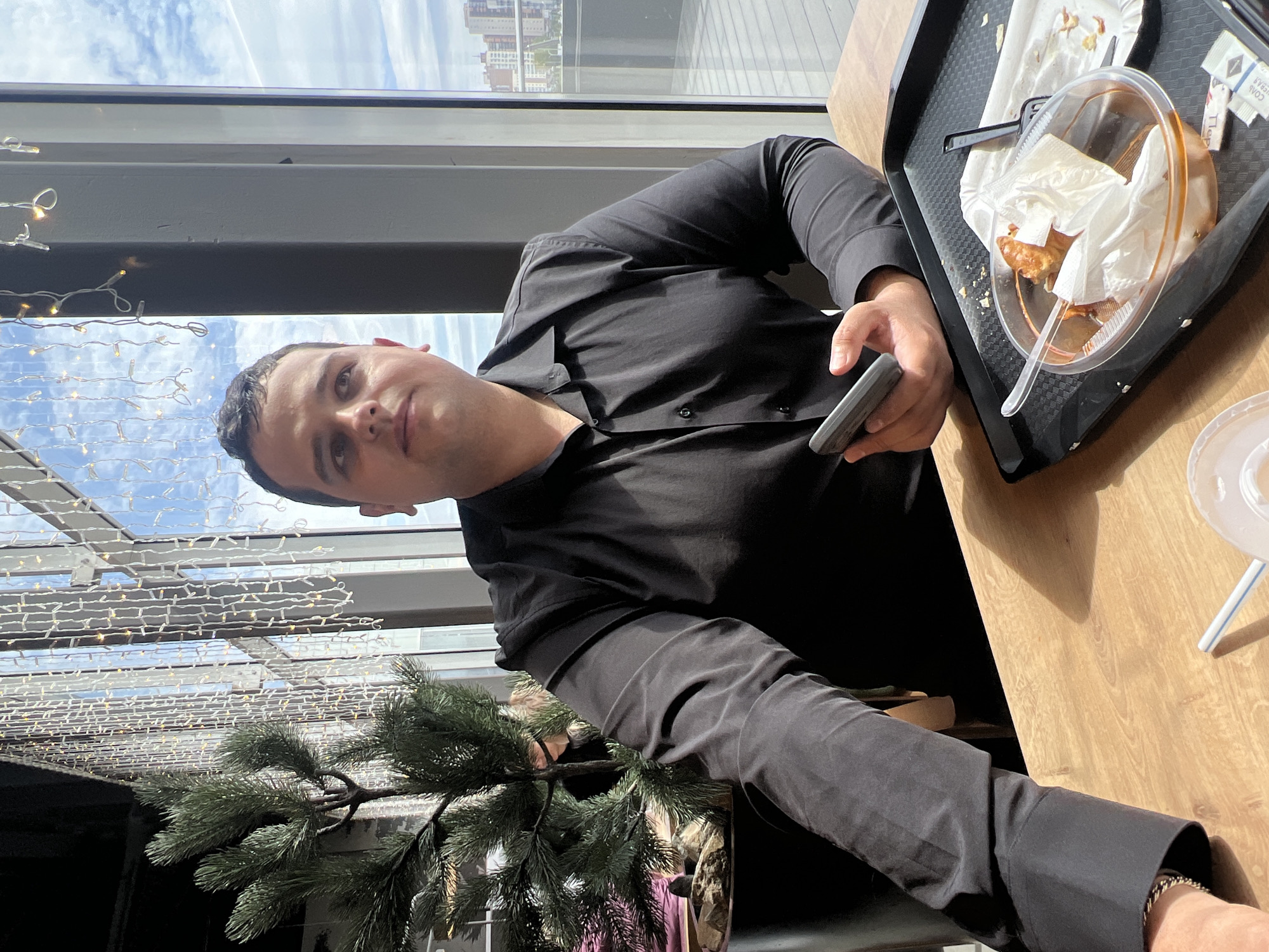 A medium shot shows a man seated at a wooden table, wearing a black, button-down shirt, and holding a grey smartphone in his right hand. He is looking towards the camera with a neutral expression. On the table to his right is a black tray with a partially consumed meal in a clear plastic container, along with a white straw and disposable condiments.
Behind the man and to his left is a window, offering a view of the sky and buildings in the city of Krasnoyarsk, Russia. The window area is decorated with strings of small lights and a small artificial tree. The light suggests a bright, daytime setting. The scene appears to be indoors, possibly in a restaurant or cafe.
