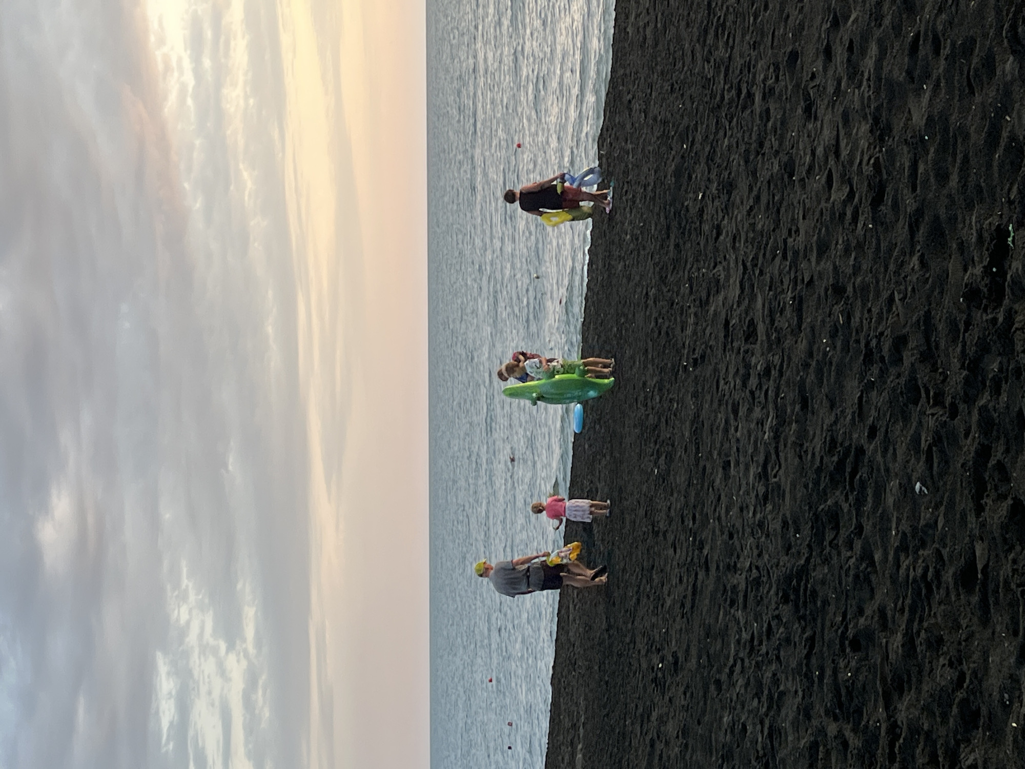 In Ureki, Georgia, on a black sand beach at dusk, several people are gathered near the calm, dark blue sea. A man in a grey t-shirt and dark shorts, wearing a yellow cap, walks across the sand holding a yellow bag. Next to him, a young girl in a pink shirt and a white skirt stands facing the sea. Further down the beach, a green inflatable toy shaped like a dinosaur is on the sand, and to its right, another person in a dark shirt and red shorts walks with several inflatable rings. The sky is filled with soft, muted clouds with hints of pink and yellow from the setting sun. The black sand beach is textured with footprints and small indentations.