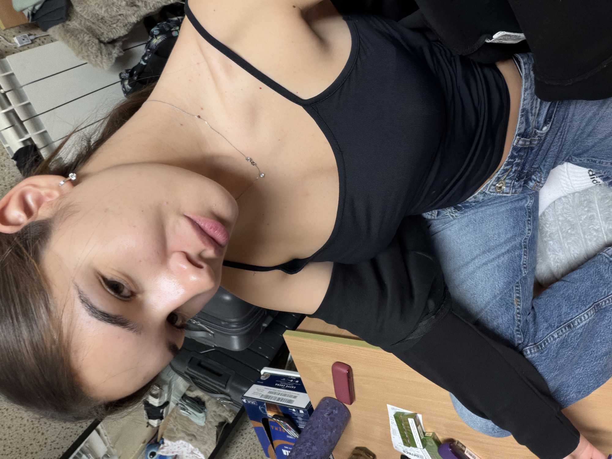 A young woman with light skin, long brown hair, wearing a black tank top and blue jeans, lies on a grey and white patterned surface, likely a bed or couch. Her head is tilted to the left. She is wearing a delicate silver necklace and a stud earring. To her left is a dark suitcase on a metal frame, partially obscured by a wooden table. On the table are several objects: a box of what appear to be small electronics (likely IQOS), a purple tube, a maroon vaping device, and some packaging from Datecs, a company that manufactures cash registers and point of sale systems. The general setting appears to be a domestic interior. There is no information to confirm this is Sofia, Bulgaria.