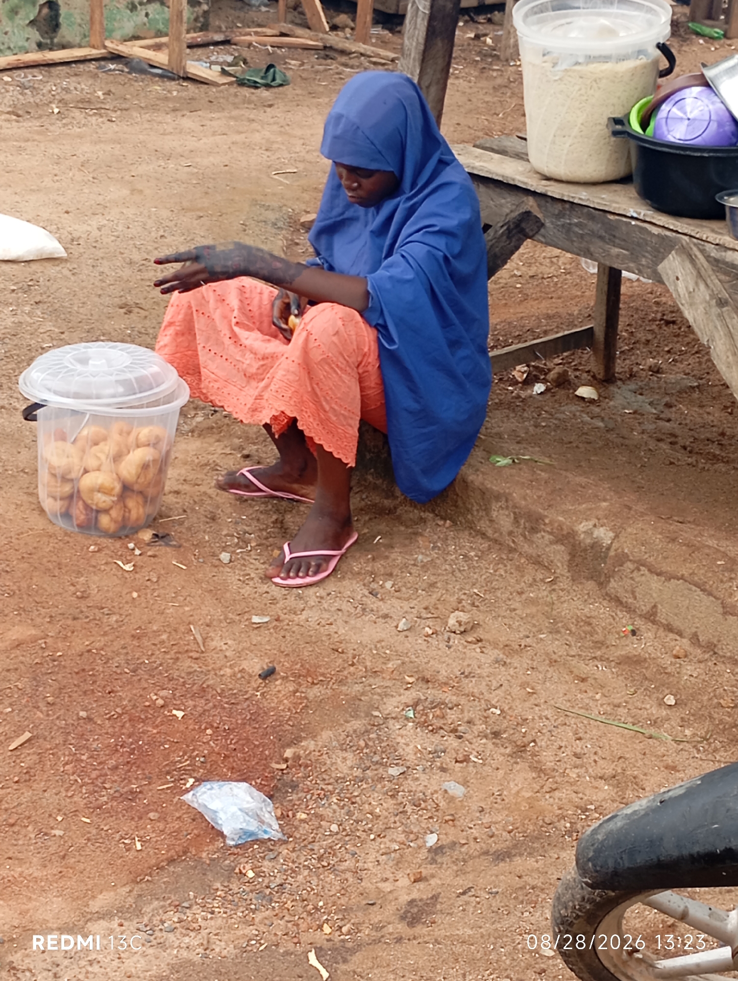 A full shot shows a young woman with dark skin wearing a blue hijab and an orange patterned skirt, sitting on a dirt embankment. Her left hand, with dark markings on the back, rests on her lap. She appears to be holding a small object. A clear plastic bucket filled with food items, possibly baked goods, sits to her left. Behind her, a wooden table holds two large containers: one filled with a light-colored grain and another with various bowls, including a purple one. The ground is a mixture of sand and gravel. The background shows a rudimentary wooden structure and some green foliage. In the lower right corner, part of a motorcycle wheel is visible. The timestamp "08/28/2026 13:23" and "REDMI 13C" are present in the lower corners of the image. The scene suggests a marketplace or roadside stall setting in Shendam, Nigeria.