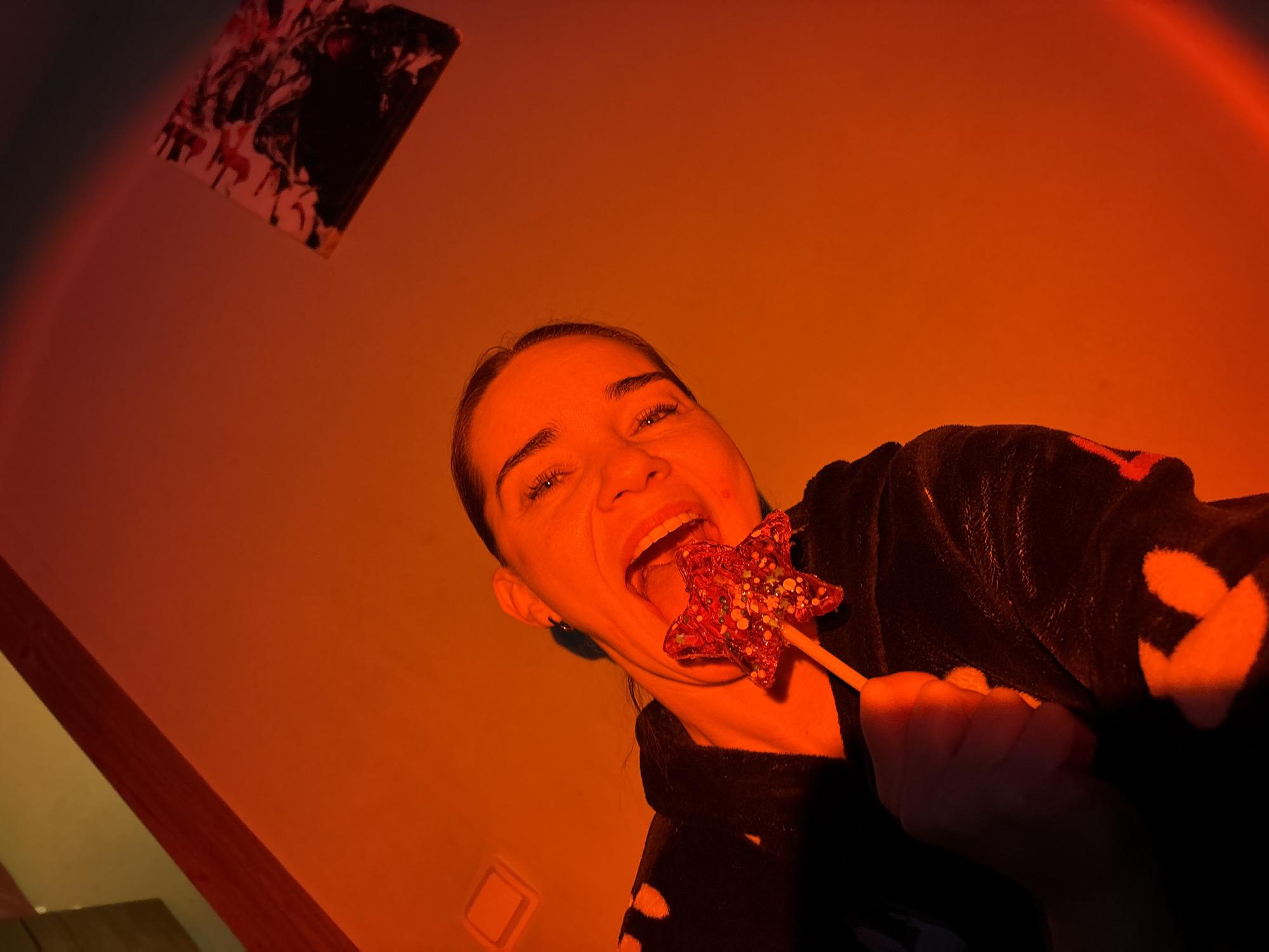 A low-angle shot shows a woman indoors with a star-shaped lollipop in her mouth. She is wearing a dark robe or jacket with light-colored, round patterns. In the background, a painting hangs on the wall. The room is illuminated with a red/orange light. The location is listed as Unknown, Russia.
