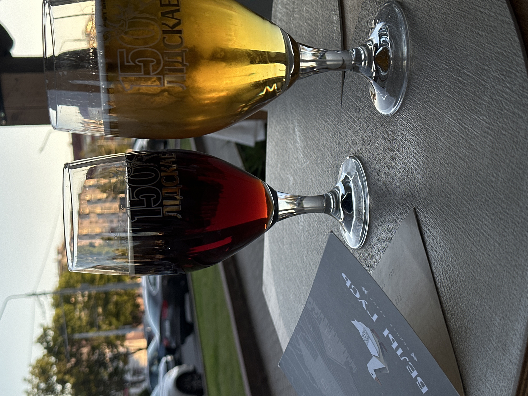 The image shows two stemmed glasses of beer on a textured table, one lighter and one darker. Both glasses have the number "150" and Cyrillic text, which appears to be "ЛІДСКАЕ," etched onto them. The lighter beer is golden yellow, and the darker beer is a rich amber. The glasses are positioned at an angle, with the stems pointing towards the lower right of the frame. A dark-colored brochure or menu is partially visible beneath the lower glass. In the background, blurred trees, cars, and some buildings suggest an outdoor setting during the daytime. The lighting indicates it is likely late afternoon or early evening, with soft sunlight casting reflections on the glasses. The overall scene appears to be a relaxed outdoor seating area, possibly at a cafe or restaurant. The text "БЕЛЫ ГУСЬ" (Bely Gus, meaning White Goose) is visible on the brochure, along with a stylized image of a goose. Based on the context provided, this scene is set in Minsk, Belarus.