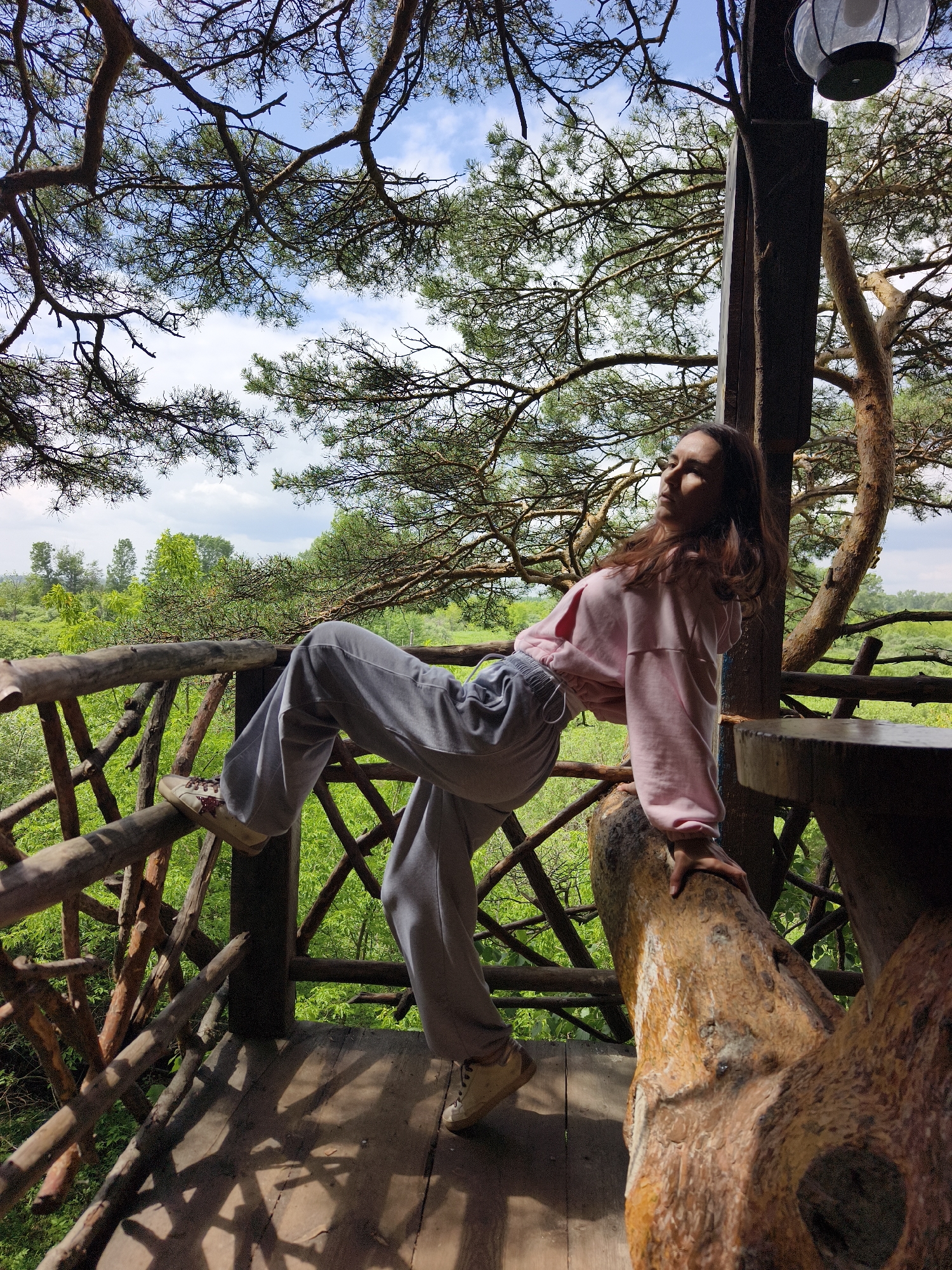 A young woman with long brown hair is posing on a wooden platform of a treehouse, leaning over a rustic wooden railing. She is wearing a pink sweatshirt and grey sweatpants. The treehouse is surrounded by lush green foliage and tall trees, with a bright blue sky and scattered clouds visible above. The platform is made of rough wooden planks, and the railing is constructed from natural branches and logs. Sunlight casts shadows across the wooden floor. The background shows a dense forest stretching out to the horizon. The overall scene conveys a sense of being immersed in nature.