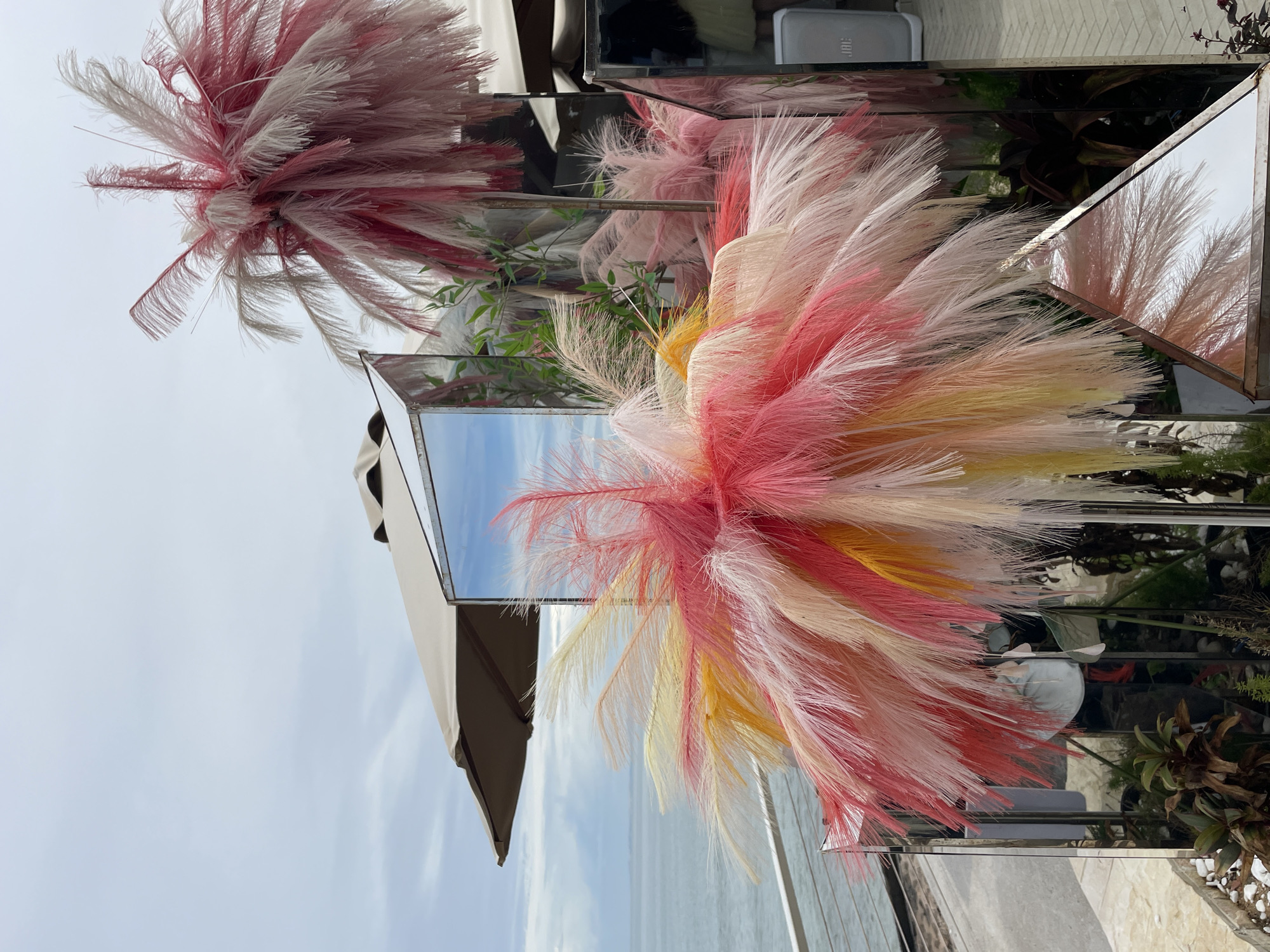 The image shows outdoor decorations in Phu Thuy Ward, Vietnam. The decorations include large, fluffy plumes in pink, white, and yellow, arranged in mirrored boxes or frames. A partially visible open umbrella is present. The sky is light blue, and a hint of the ocean can be seen in the reflections. Plants and foliage are visible within the mirrored structures. A JBL speaker is on top of one of the mirrored boxes.