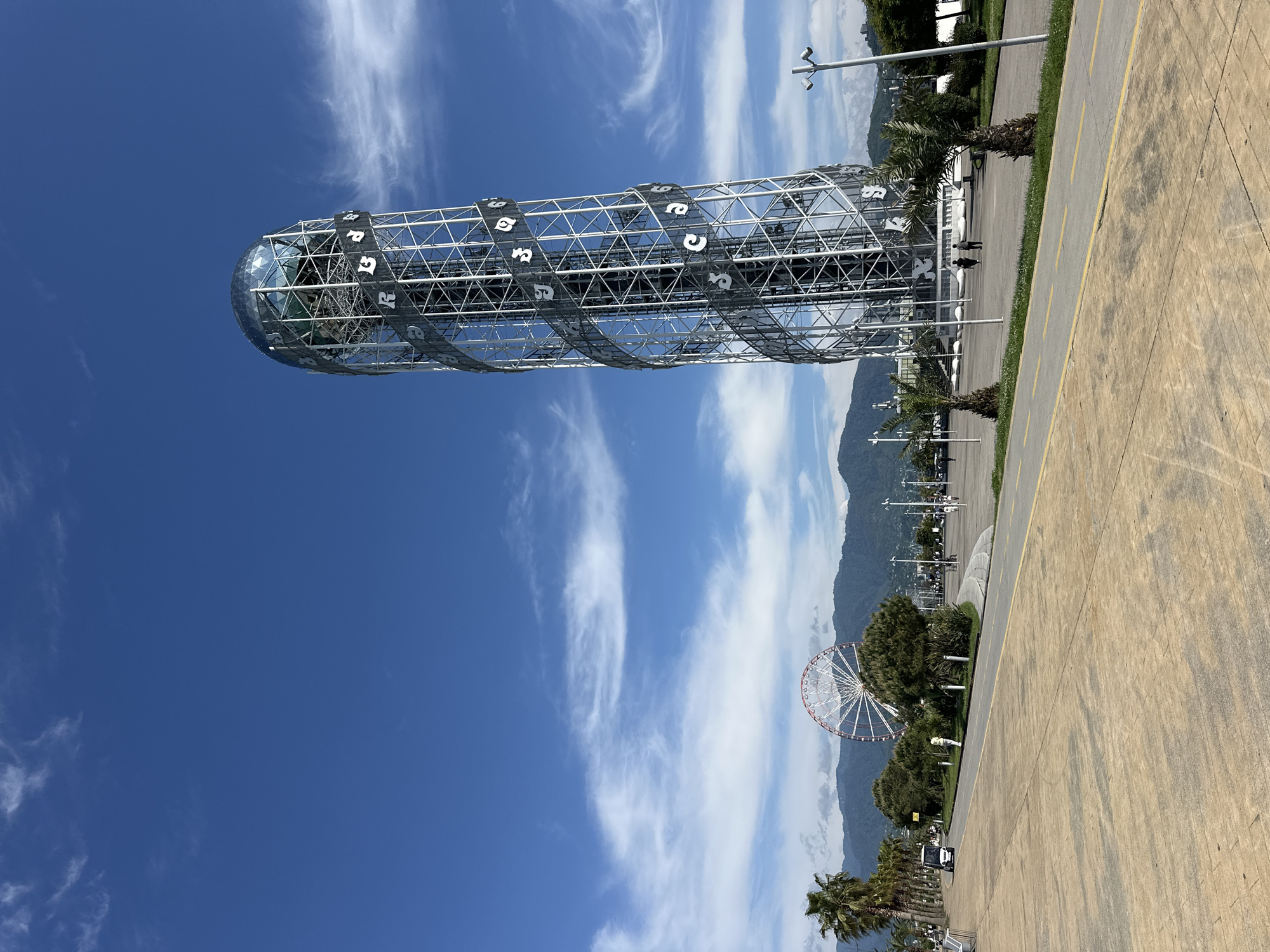 The image shows a vertical view of the Alphabet Tower, a 130-meter tall monument in Batumi, Georgia. The tower's unique DNA-shaped structure is visible, featuring Georgian alphabet letters. A Ferris wheel can be seen in the background. The sky is bright blue with scattered clouds. Paved walkways and palm trees are also visible.