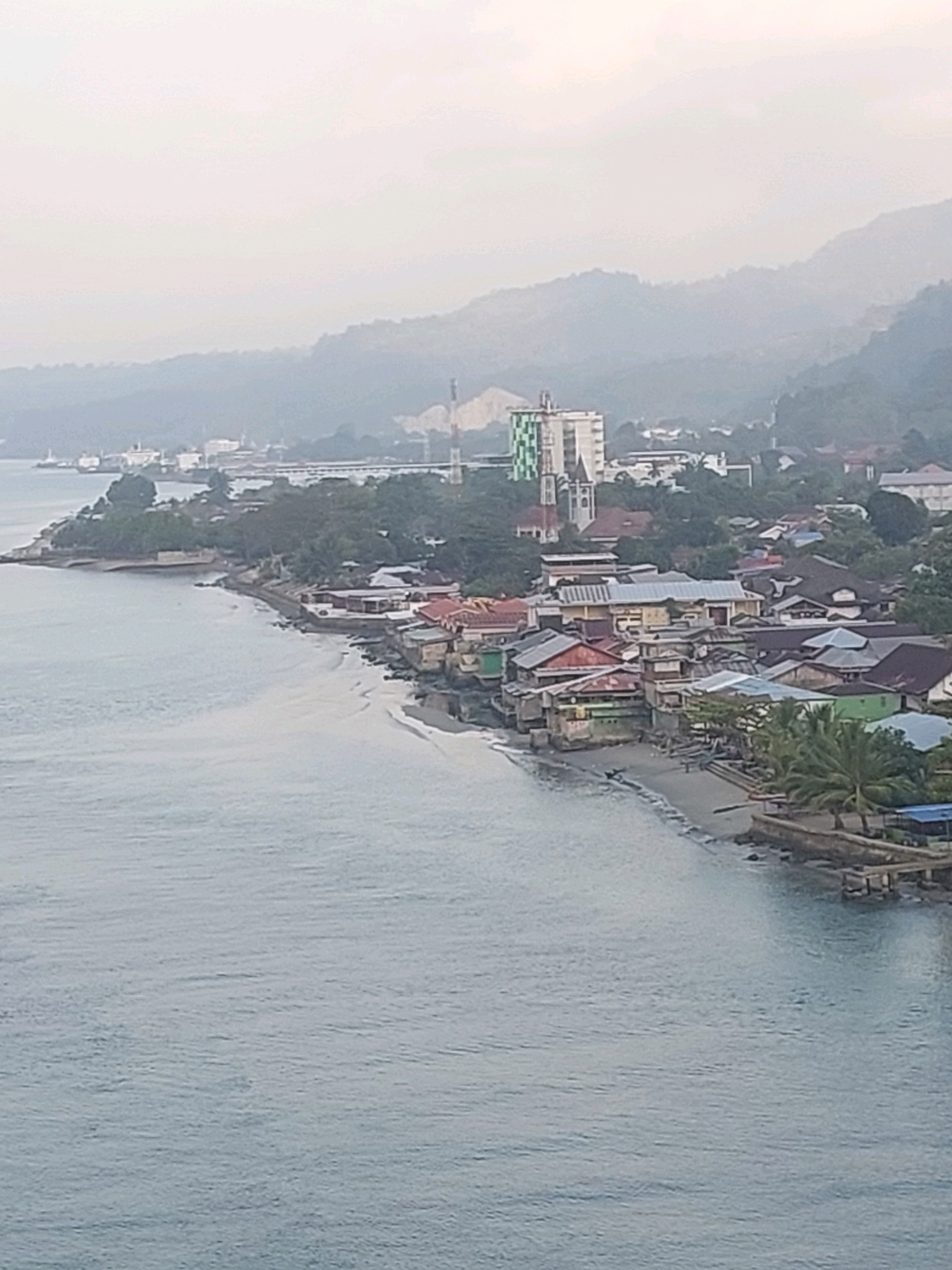 The image displays a coastal town with a dense collection of buildings situated along a shoreline. The architecture varies, with some structures appearing to be houses with colorful roofs, while others are larger, more modern buildings. A tall building with a green and white checkered facade stands out among the others. The town is nestled against a backdrop of lush, hilly terrain that fades into a hazy sky. In the foreground, the calm, gray water of the sea laps at the sandy beach. There are some boats visible in the distance near a port or industrial area. The scene suggests a relatively overcast day. The location appears to be the coastal area of Ambon, Indonesia, likely facing a bay or harbor given the presence of port infrastructure. There are no visible people or specific actions taking place. The focus is on the urban landscape and its relationship with the natural environment.