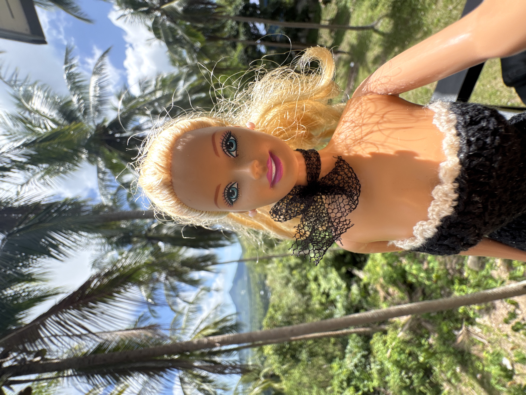 In a close-up, frontal view, a blonde doll with blue eyes and bright pink lips is positioned against a backdrop of lush greenery and palm trees under a partly cloudy blue sky. The doll is wearing a black crocheted top with a white trim. A black lace accessory is visible around its neck. The background suggests a tropical outdoor setting, possibly a hillside overlooking a landscape with a distant mountain. The lighting indicates it is daytime, casting shadows on the doll's shoulder and arm. The scene is captured from a low angle, placing the doll prominently in the foreground. The location is identified as Ban Tai Subdistrict, Thailand.