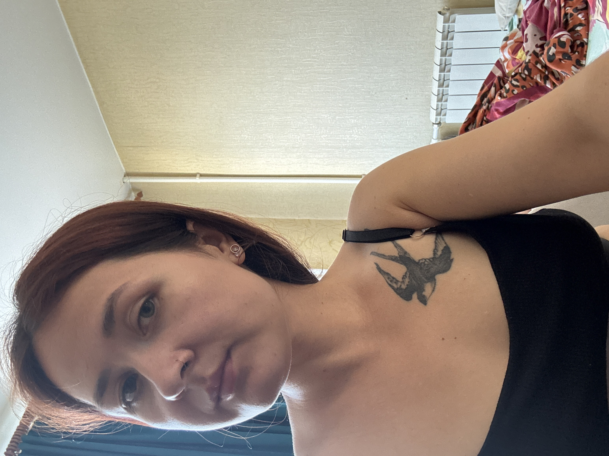 A young woman with auburn hair and fair skin is shown in a medium close-up, posing for a selfie. She is wearing a black tank top, and a tattoo of a bird in flight is visible on her left collarbone. She has a nose ring and small hoop earrings. The background consists of a plain, textured yellow wall and a white radiator. The lighting suggests an indoor setting during the daytime. The image is a personal photograph, likely taken in a residential environment. The exact location within Naberezhnye Chelny, Russia cannot be determined from the visual information alone.