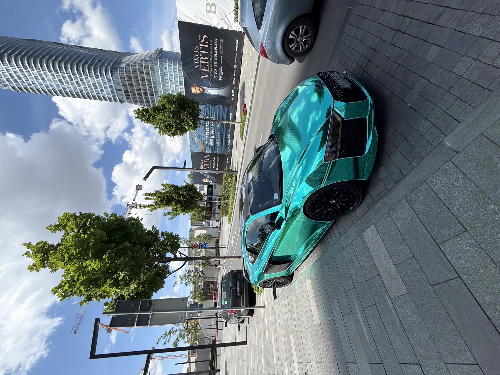 The image depicts an urban scene in Belgrade, Serbia. Dominating the left side is a towering, modern skyscraper against a partly cloudy sky. Adjacent to the skyscraper, a large billboard advertises an event called "Nikos Vertis" and "Belgrade River Fest."
A strikingly colored, turquoise car is parked alongside the curb. A dark-colored car with the license plate "PA 152-LV" is parked behind it. The foreground consists of a tiled sidewalk made of stone paving blocks. Trees line the street and construction cranes are visible in the background.