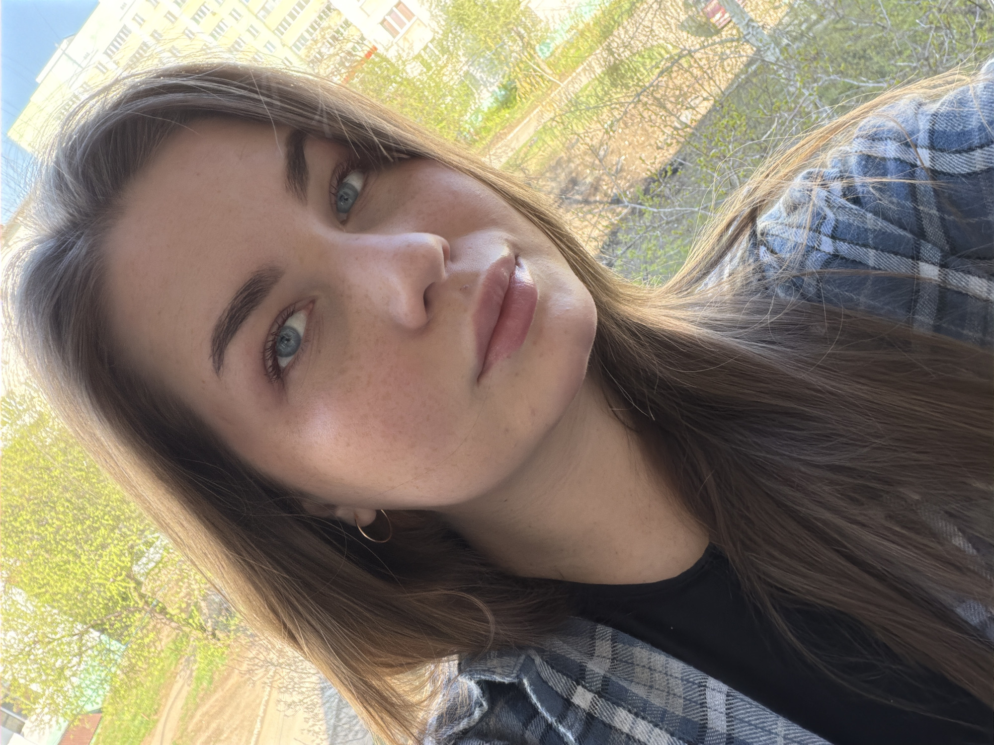 The image depicts a young woman with light skin, freckles, blue eyes, and brown hair. She is wearing a blue and grey plaid shirt over a black top. The background shows trees and buildings, suggesting an urban environment. The picture was taken in Chaikovsky, Russia.