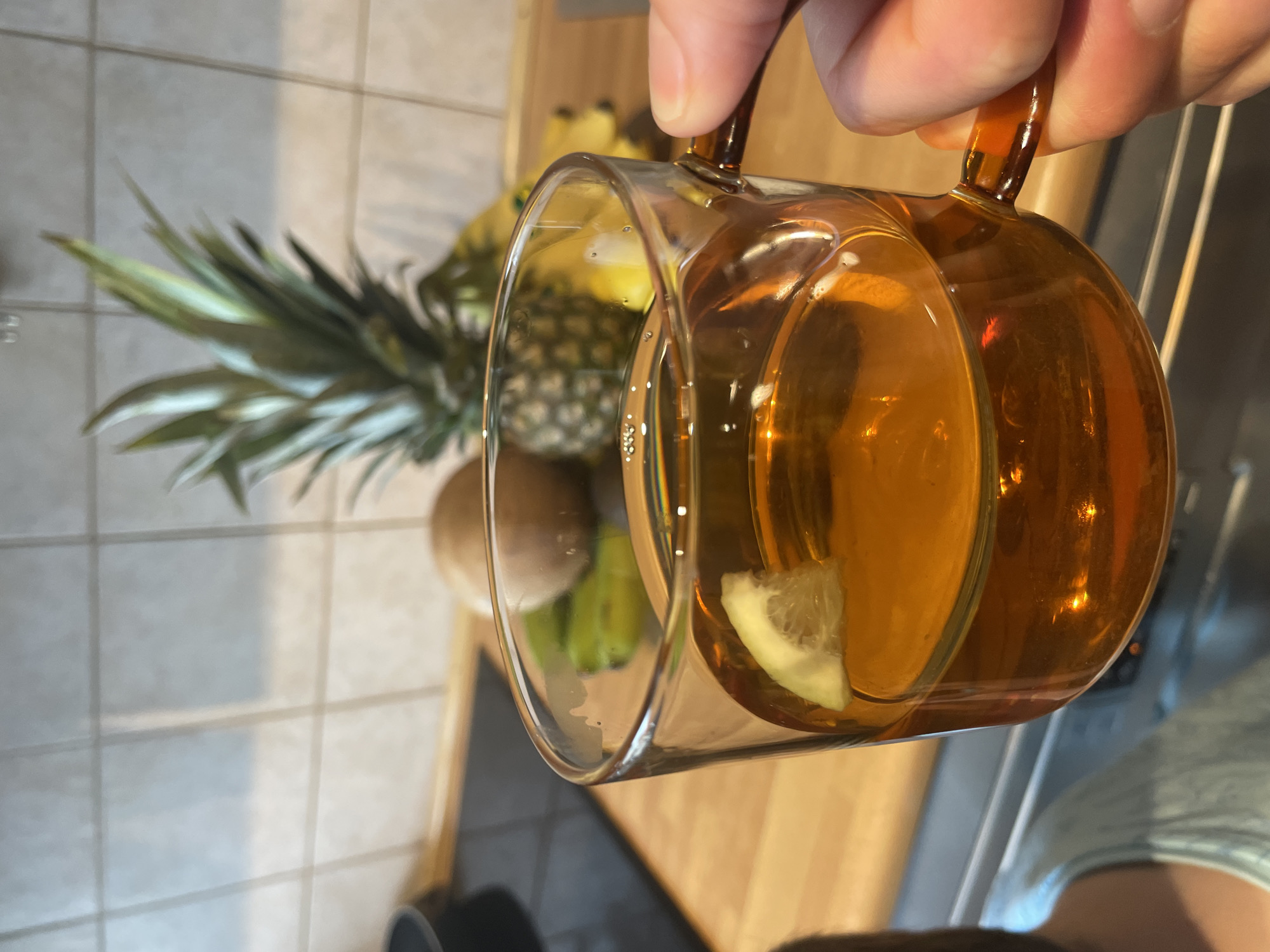 A close-up shot shows a hand holding a clear glass mug filled with amber-colored liquid and a slice of lemon. In the background, a pineapple, bananas, and what appears to be a kiwi sit on a wooden countertop. The kitchen wall behind the fruit is tiled. The scene is indoors.