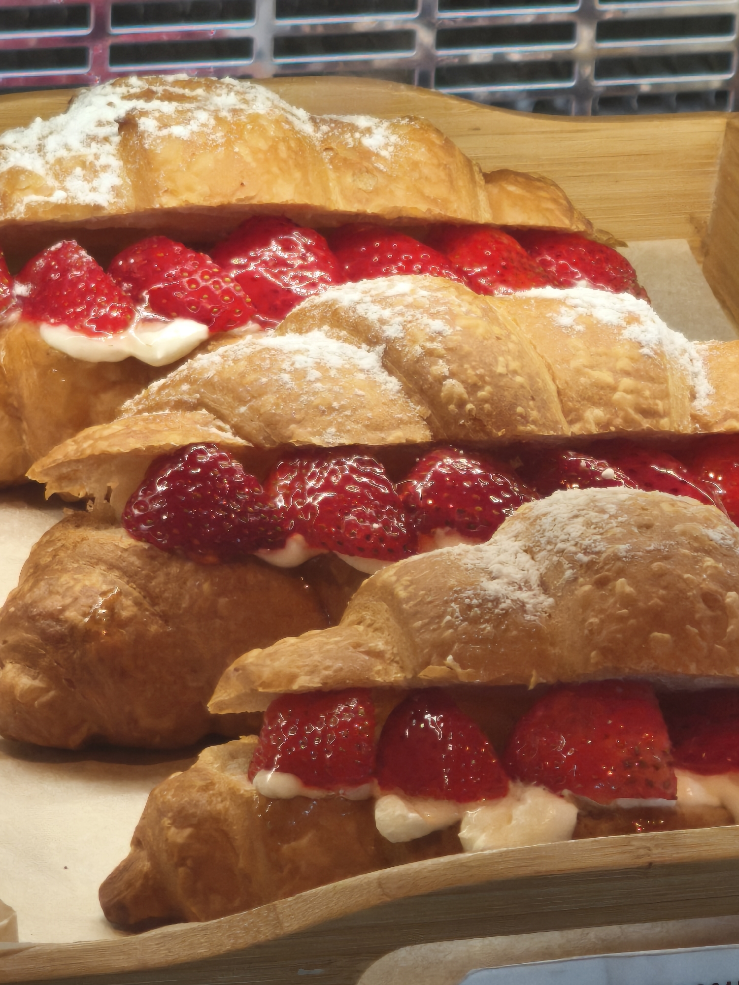 A close-up, high-angle shot shows three stacked strawberry cream croissants on a wooden tray. The croissants are dusted with powdered sugar and filled with fresh strawberries and cream. The background appears to be a display case, suggesting these pastries are for sale in a bakery or patisserie. The lighting is bright, highlighting the textures and colors of the food. No people or specific city details are visible in the image.