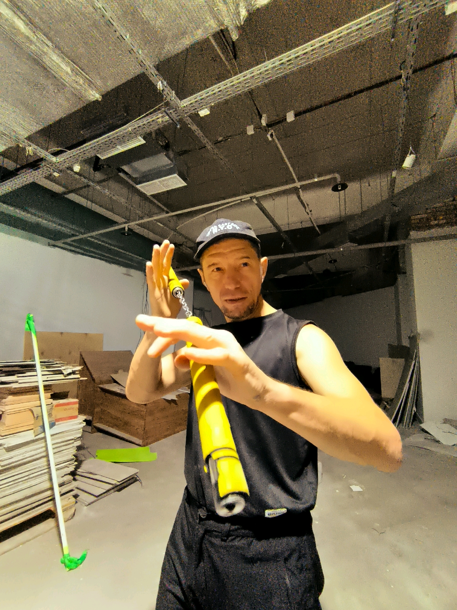 A man in a black sleeveless shirt and black pants, wearing a black baseball cap, stands in an unfinished room holding yellow nunchucks. He is looking towards the camera with his mouth slightly open. The room has a concrete ceiling with exposed pipes and conduits, and bare concrete walls. Piles of wood, cardboard, and other materials are stacked in the background. A green-headed broom stands leaning against a pile of materials on the left side of the image. The floor is concrete. The lighting is dim and appears to be from overhead fixtures.