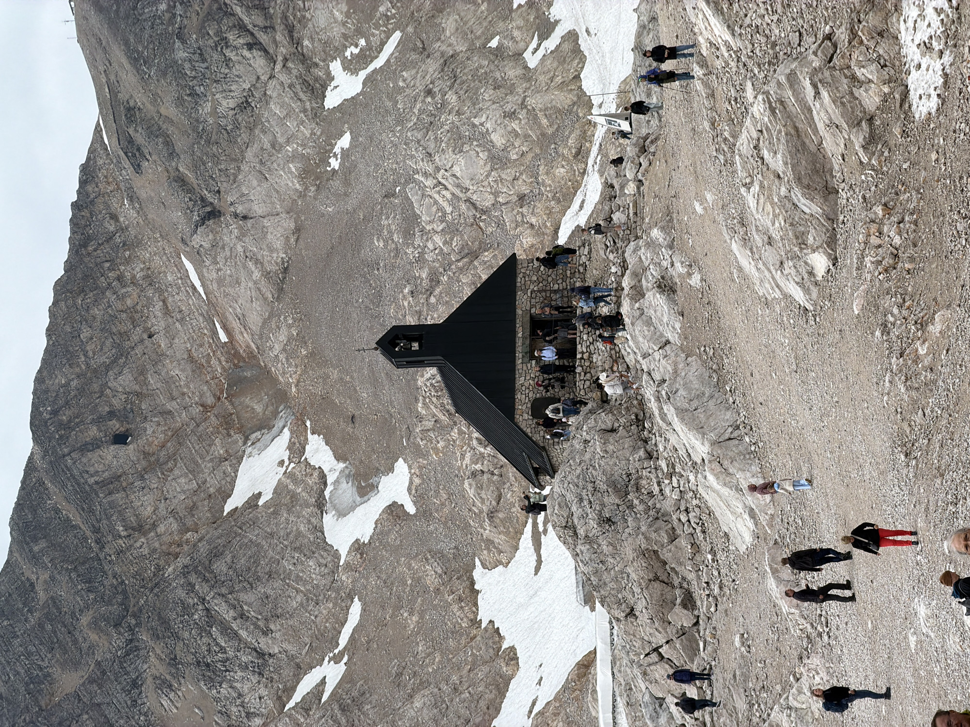 The image depicts the view from the Zugspitze, Germany's highest peak, located near Garmisch-Partenkirchen. A small, dark-roofed building, possibly a chapel or restaurant, sits prominently on a rocky, barren mountainside dotted with patches of snow. A group of tourists are visible, some gathered near the building, others walking on the rocky terrain. The surrounding landscape is rugged and mountainous, with steep, textured rock faces and a cloudy sky.