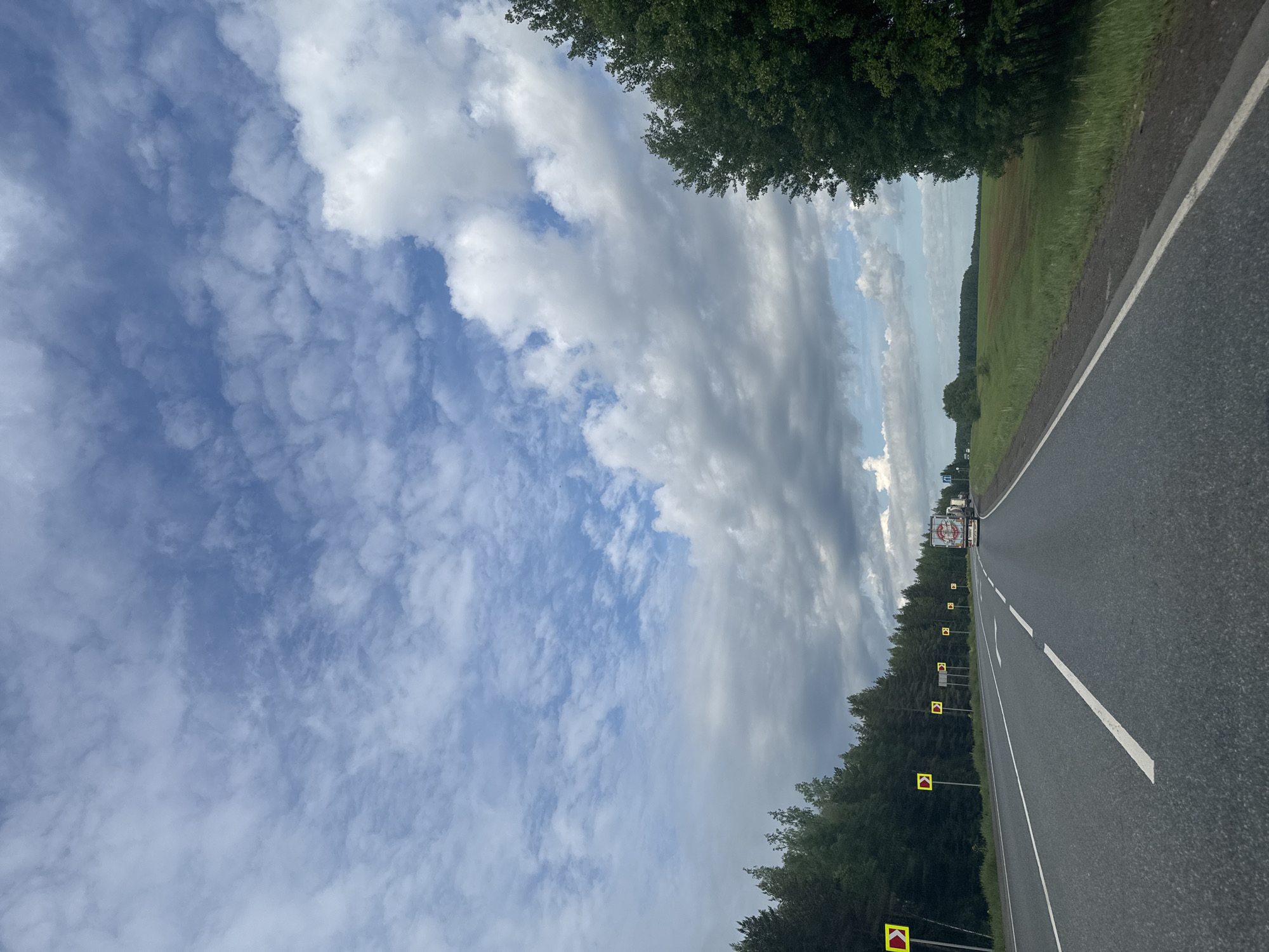 An image, rotated 90 degrees clockwise, captures a view along a rural road, likely from a moving vehicle, under a partially cloudy sky. The asphalt road occupies the right side of the frame, featuring a solid white line on its outer edge and a broken white line separating lanes, indicating a two-lane highway. To the left of the road, a dense line of green trees runs parallel, acting as a border to a vast open field. The field, visible beyond the treeline in the upper right, displays shades of green and brown, suggesting agricultural land.
Numerous yellow and red chevron road signs are spaced along the left side of the road, indicating a curve or hazard ahead. In the distance, a large white truck is visible traveling in the same direction. The sky above is a mix of blue patches and extensive cloud cover, ranging from fluffy white cumulus clouds to darker, more spread-out gray clouds. The lighting suggests daytime, possibly mid-day or late afternoon. No people are visible. While there is text on the distant truck, it is too small and blurred to be legible. The setting is consistent with a countryside road in Dybki, Russia.
