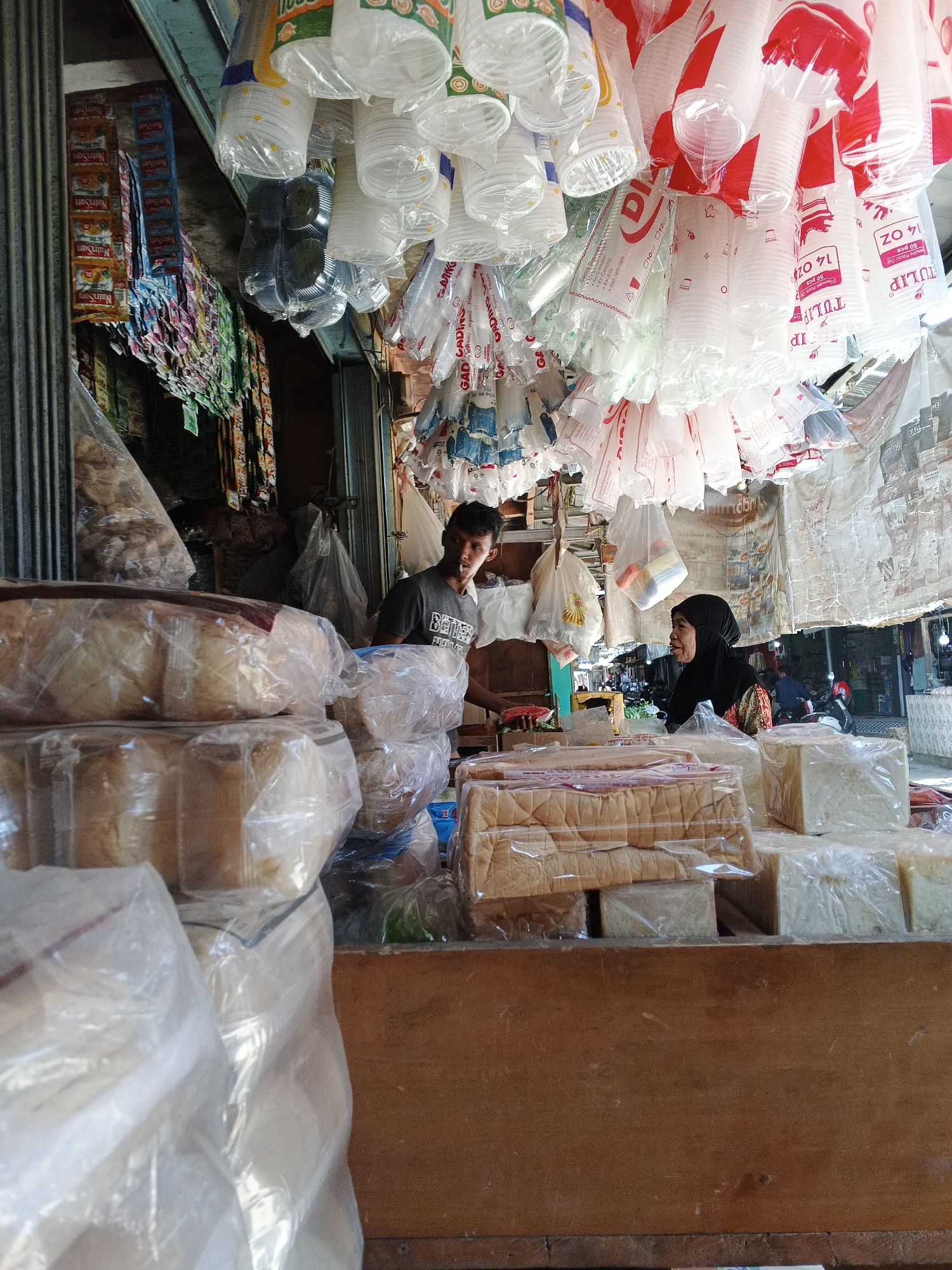 In Warungkiara, Indonesia, a small market stall is captured with a man and a woman present. The stall displays an array of goods, with stacks of bread wrapped in plastic prominently featured in the foreground. Overhead, numerous plastic cups hang in bunches, some red and white, others clear. To the left, rows of packaged snacks or sachets are suspended. The man, wearing a dark t-shirt, stands behind the counter. The woman, dressed in a headscarf and patterned clothing, is positioned further back in the stall. The overall impression is of a busy, local marketplace setting.