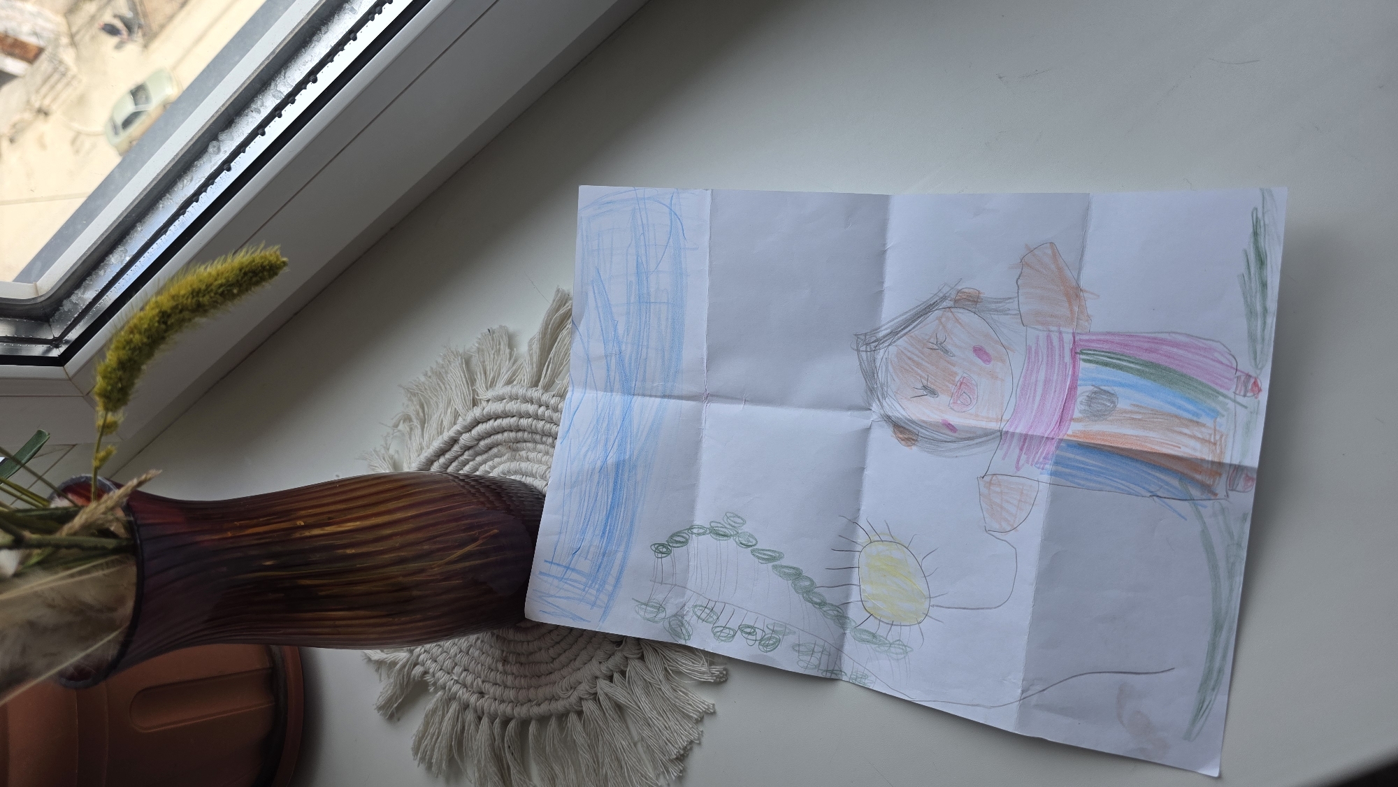 The image shows a child's drawing on a folded sheet of paper, lying on a white windowsill in what appears to be Lensk, Russia, based on the window view. The drawing depicts a figure, possibly a person, with a colorful shirt, a sun, and blue scribbles that could represent water or the sky. The windowsill also holds a brown vase filled with dried plants, resting on a macrame mat. The overall scene has a casual, domestic feel.