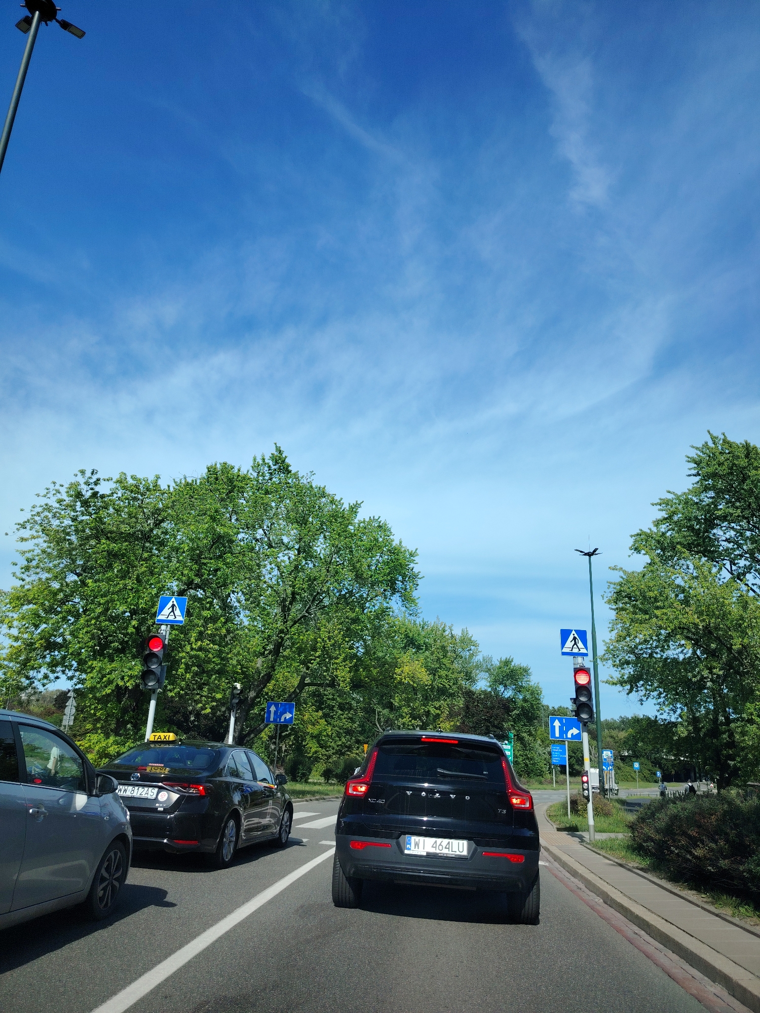 In a street scene in Warsaw, Poland, on a clear, sunny day, traffic is stopped at a red light. A grey car is in the left lane, a black taxi with a yellow "TAXI" sign is in the middle lane, and a black Volvo XC40 is in the right lane. A pedestrian crossing sign is visible next to the traffic lights. The sky is bright blue with scattered white clouds. Trees line the sides of the road, and a sidewalk is present on the right. The license plates of the vehicles are visible: WW 812AS on the taxi and WI 464LU on the Volvo.