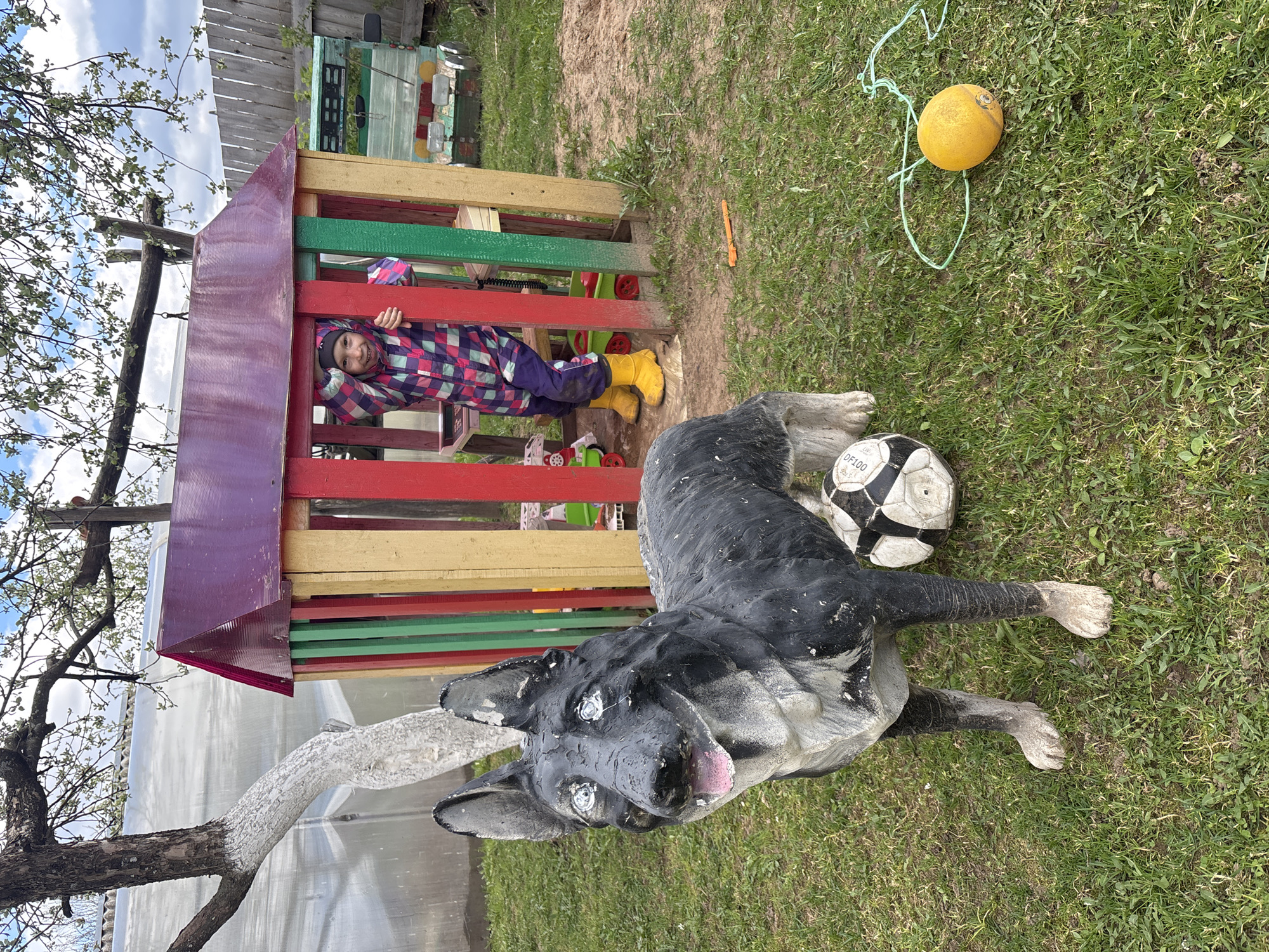 A young child in a colorful plaid jumpsuit and yellow boots peeks out from a colorful playhouse in a grassy area. A large, cement dog statue lies on the grass next to a soccer ball. A yellow ball with a blue string sits further in the grass. The location appears to be in a yard in Ваньки, Russia.