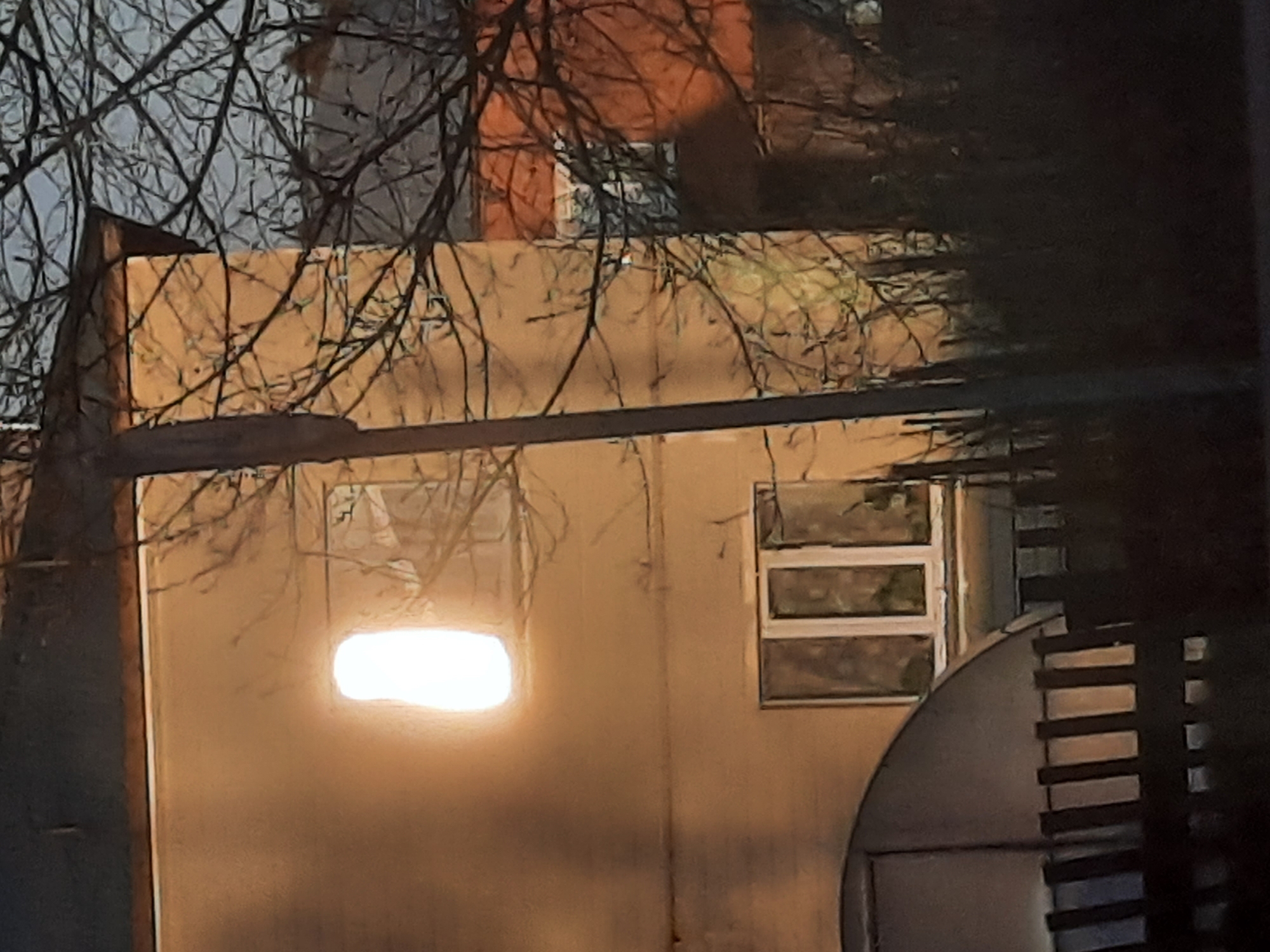 The image displays a nighttime scene featuring a light-colored building, likely a single-story structure, partially veiled by a dense network of bare tree branches. The building's facade is a muted beige or pale yellow. On the left side, a prominent, bright oblong light illuminates from within, casting a strong glow on the exterior wall around what appears to be a window. To the right of this light, another window with a white frame and multiple rectangular panes is visible.
Above the main building, glimpses of an orange or reddish structure, possibly another building or a roof, can be seen through the branches against a dark sky. On the far right, a dark, curved element with vertical slats or rungs is partially visible, which could be part of a fence, gate, or an architectural detail of an adjacent structure. The bare branches suggest a dormant season, such as winter or late autumn. There are no people visible, and no specific activity is taking place other than the illumination from inside the building. No visible text is present. The setting is Krasnogorskoye, Russia.