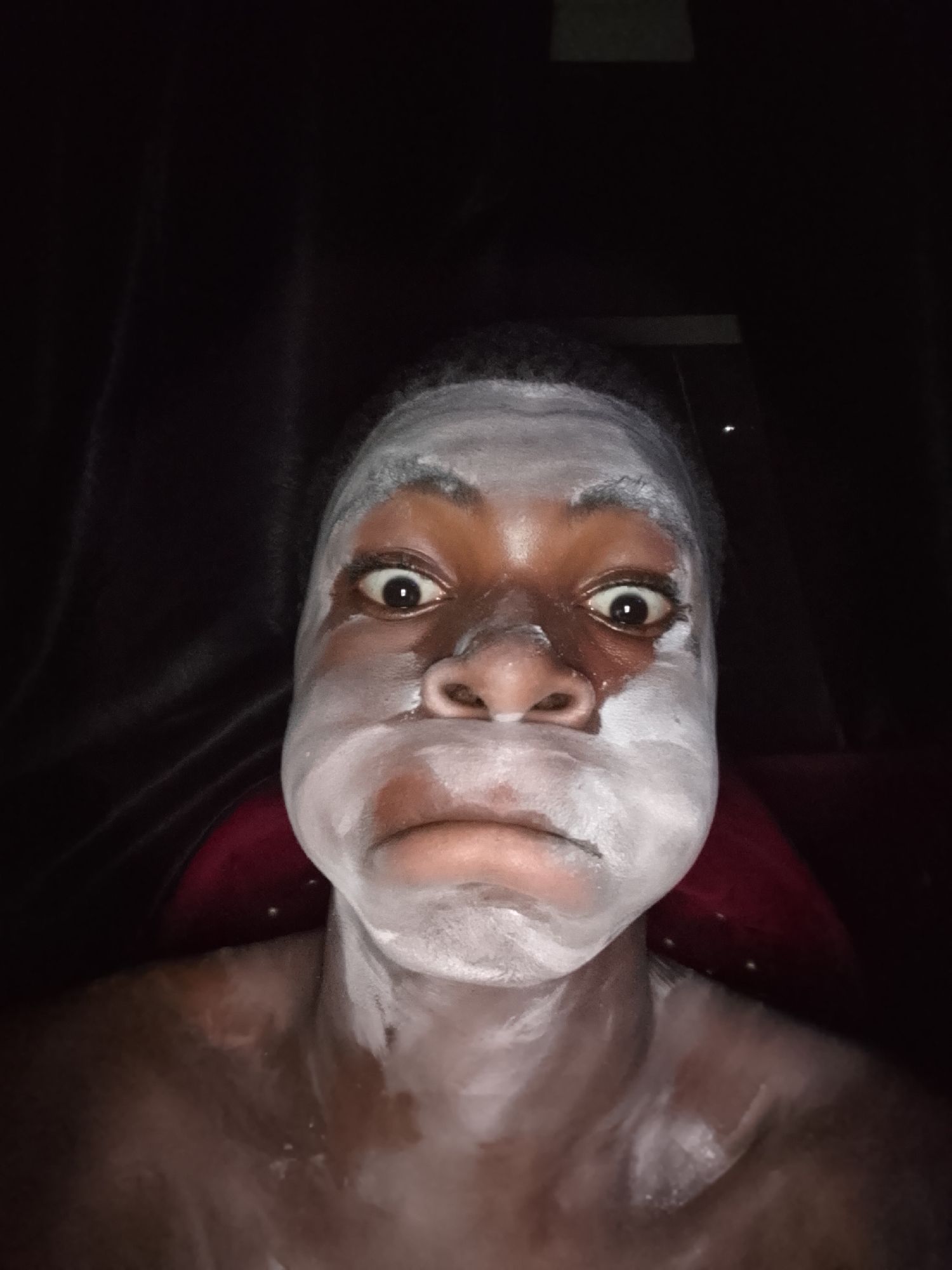 A dimly lit, close-up portrait features a person with dark skin, whose face and upper body are extensively covered in a white, pasty substance. The subject looks directly at the camera with wide, exaggerated eyes, their pupils dilated. Their mouth is slightly pursed in a frown or pout. The white substance, resembling a face mask or paint, is applied unevenly, thickest on the cheeks and forehead, with streaks and drips visible on the bare shoulders and chest. The background is very dark, hinting at an indoor setting with low lighting, possibly with dark curtains or drapes behind the person. A subtle reddish color can be seen behind the subject's right shoulder, perhaps from clothing or a seat. No text is visible in the image.