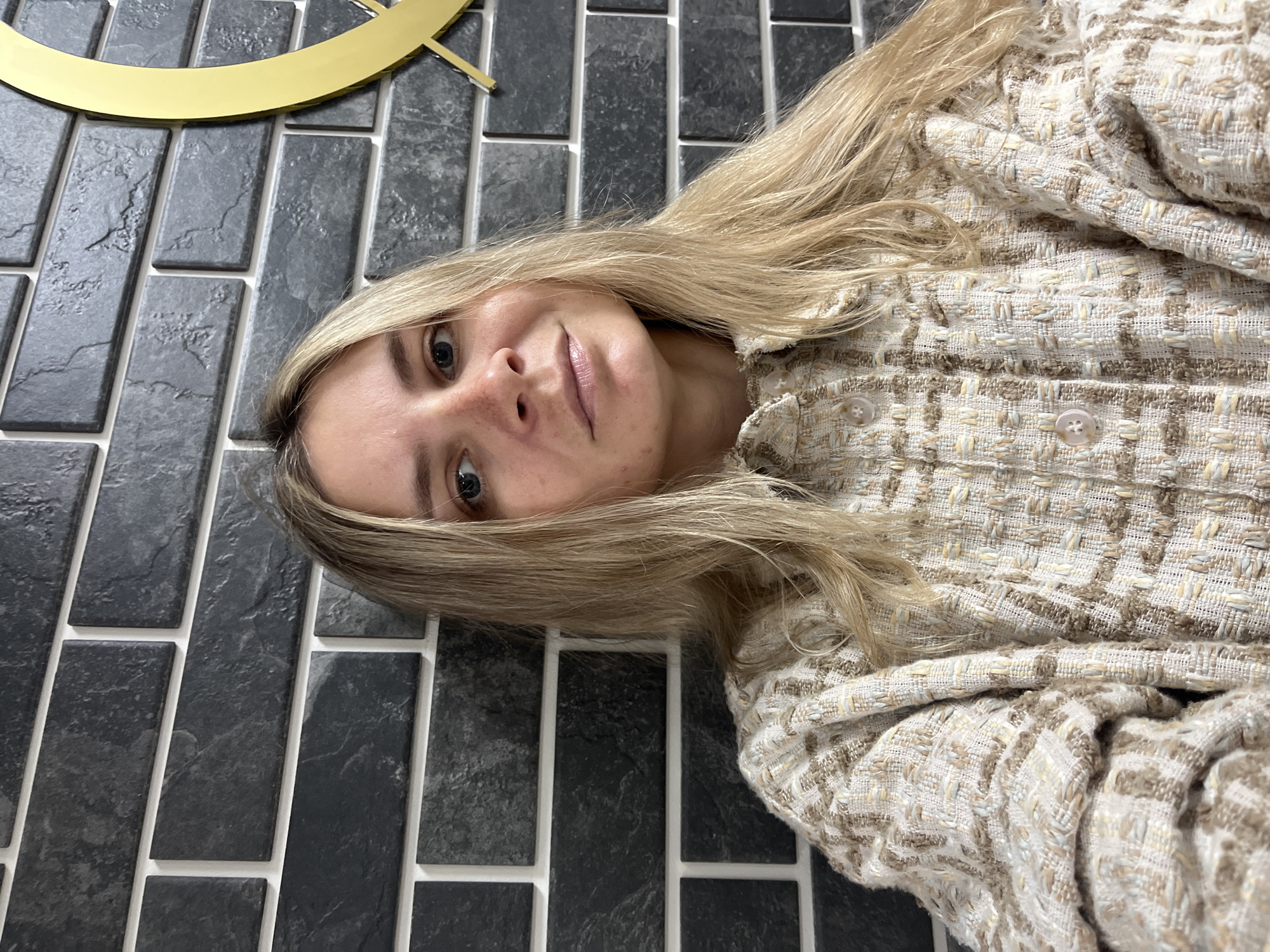 A portrait-style image, rotated 90 degrees clockwise, features a young woman with long, straight blonde hair lying on a dark-tiled surface in what appears to be an indoor setting. She has fair skin, blue/grey eyes, and a neutral expression with a hint of a smile, looking directly at the camera. A few freckles are visible on her nose and cheeks.
She is wearing a textured jacket, possibly tweed, in shades of light beige, cream, and brown, with a visible woven pattern and small light-colored buttons down the front. Her hair is spread around her head and shoulders, blending with the tones of her jacket.
The background consists of rectangular, dark grey or black tiles with a slightly textured, stone-like appearance, laid in a brick pattern with prominent white grout lines. In the upper left corner of the frame, a curved, golden metallic object, possibly part of a large clock or abstract wall decoration, is partially visible against the dark tiles. The lighting suggests an indoor environment, with no specific time of day or weather discernible. No text is visible in the image. The setting is located in Chaikovsky, Russia.