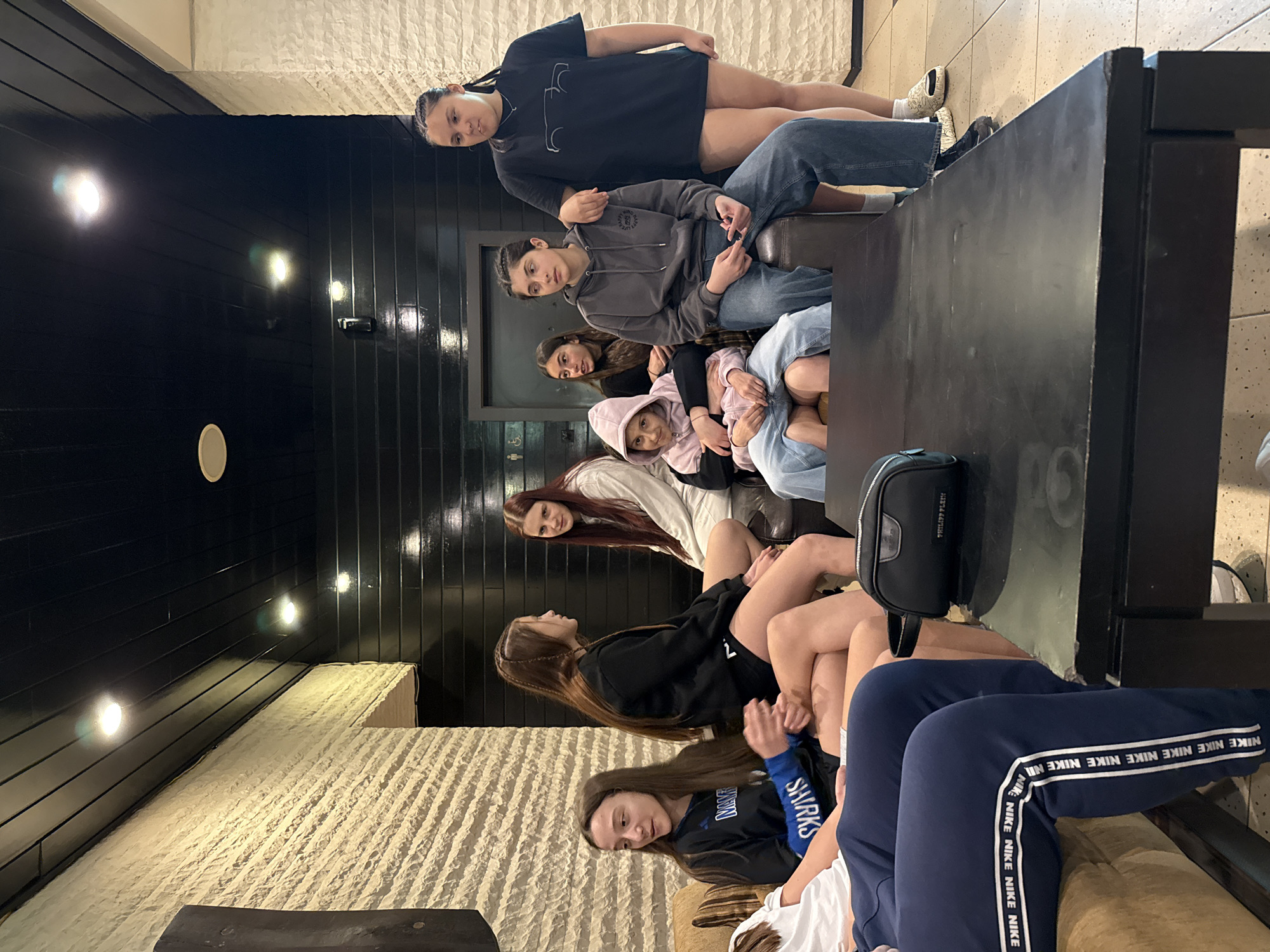 The image depicts a group of seven young women sitting on couches in what appears to be an indoor space. The room features textured, light-colored walls on one side and dark, paneled walls with inset lighting on the other. A dark, flat-topped surface, possibly a table or a wide armrest, runs along the center of the scene. The women are dressed in casual clothing, including hoodies, t-shirts, jeans, shorts, and track pants with the "NIKE" logo. They are positioned in a line, and their faces are visible. The general setting suggests a lounge or waiting area. There is a black bag resting on the table surface. The presence of the name "Etropole" and "Bulgaria" in the response is due to the request to include the location information in the description.