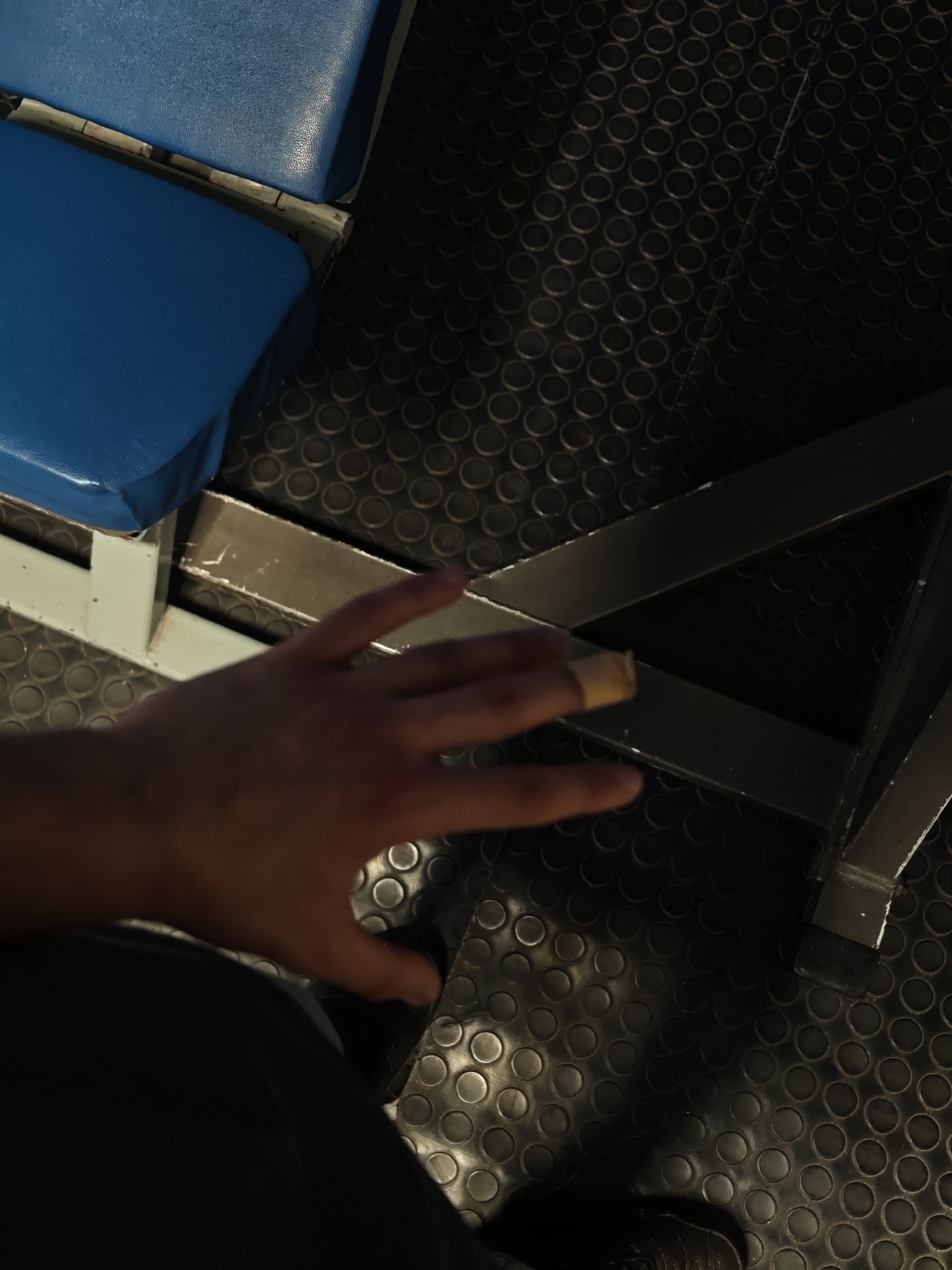 The image shows a first-person perspective of a person sitting on a blue padded seat in what appears to be a gym or fitness area. The person's left hand is visible, with a bandage on their index finger. The floor is black rubber with a circular tread pattern. In the background, there is metal support structure. The location in the city of Kozloduy, Bulgaria cannot be determined from this image alone.