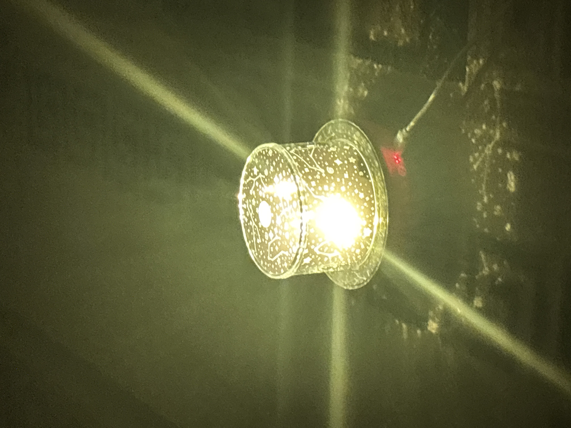 The image depicts a cylindrical lamp with a starry pattern, emitting a bright yellow light. The lamp sits on a circular base, which is placed on a dark, reflective surface. A cable is connected to the base of the lamp, and a small red light is visible near the connection point. Two prominent streaks of light, likely reflections or beams, cut across the scene diagonally from the upper left to the lower right and from the upper right to the lower left. The background is dark and appears to be a wall or surface with some texture and light variations. The scene is likely indoors, possibly in a dimly lit room. There is no indication of the location being Kryvyi Rih, Ukraine.